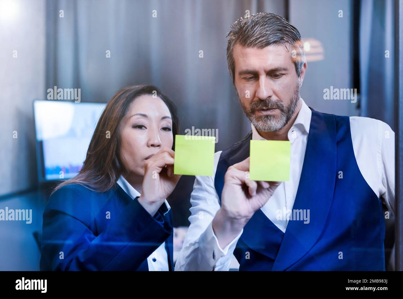Uomo adulto serio e donna in abiti formali in piedi vicino alla finestra dell'ufficio e la scrittura di idee sui foglietti adesivi gialli Foto Stock