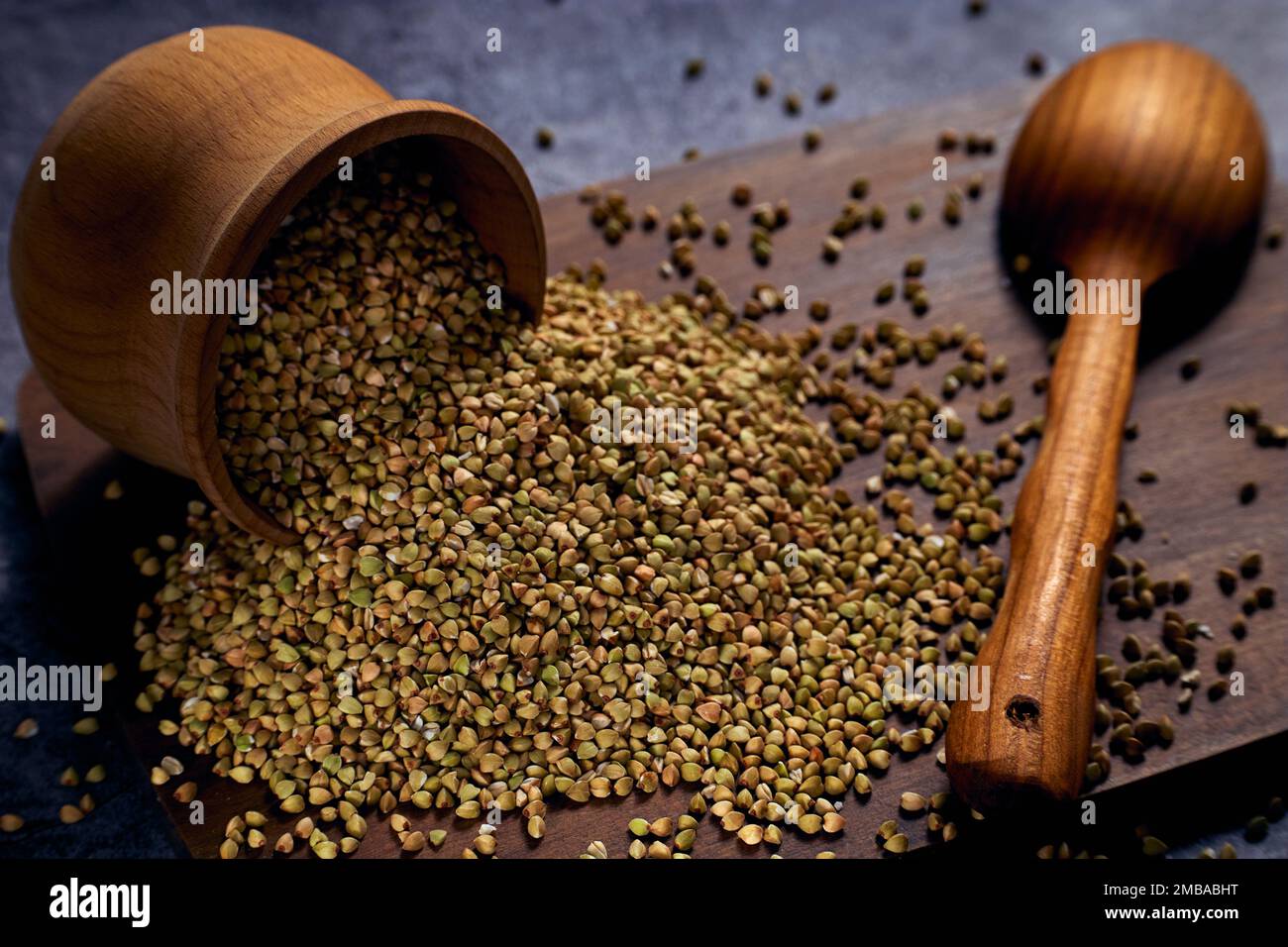 Ciotola di legno rovesciata con grano saraceno verde non lavorato su tavola di legno con un cucchiaio di legno accanto ad essa Foto Stock