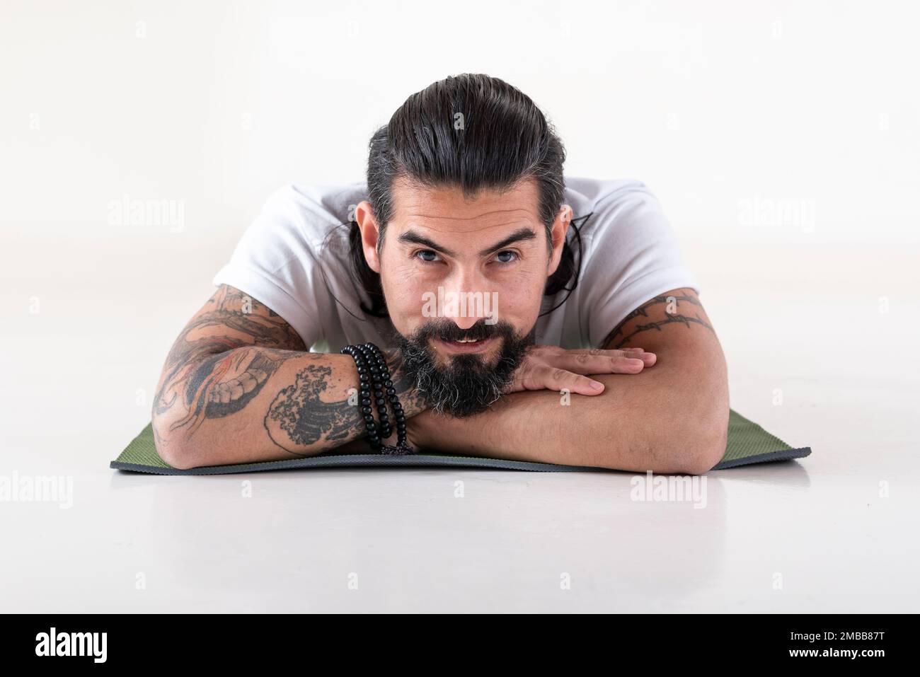 Ritratto di un uomo vestito di abiti bianchi yoga sdraiato su un tappetino mentre si guarda la fotocamera su sfondo bianco. Foto da studio. Foto Stock