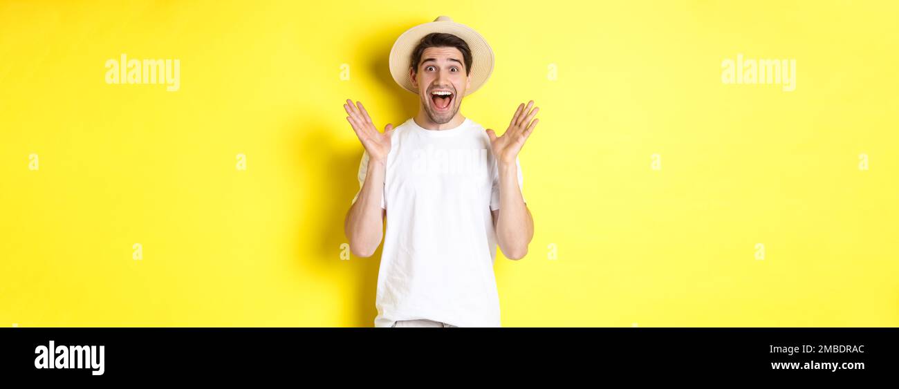 Concetto di turismo ed estate. Felice giovane uomo in cappello di paglia che guarda stupito, reagendo a sorpresa, in piedi su sfondo giallo Foto Stock