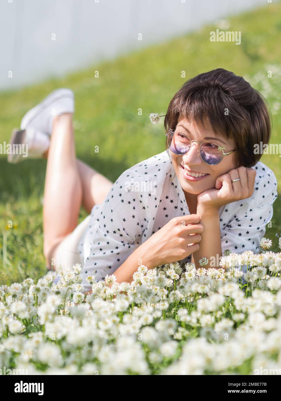 Donna in occhiali da sole colorati sniffs fiori trifoglio sul prato in parco urbano. Natura in città. Rilassati all'aperto dopo il lavoro. Atmosfera estiva. Foto Stock