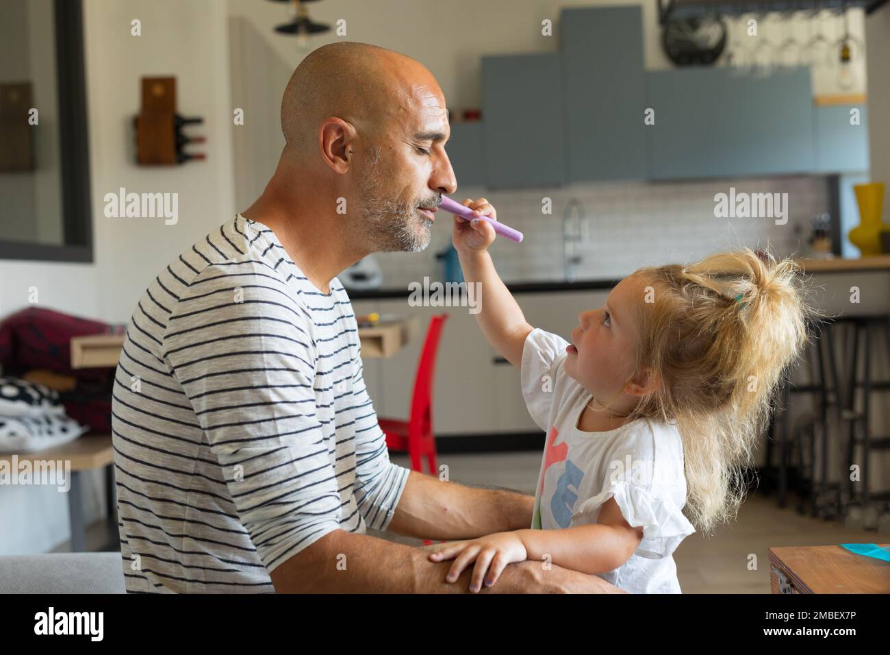 la bambina allunga il braccio per dipingere e mettere il make-up sul volto del padre; giocano il make-up nel soggiorno, il padre tiene gli occhi chiusi e. Foto Stock