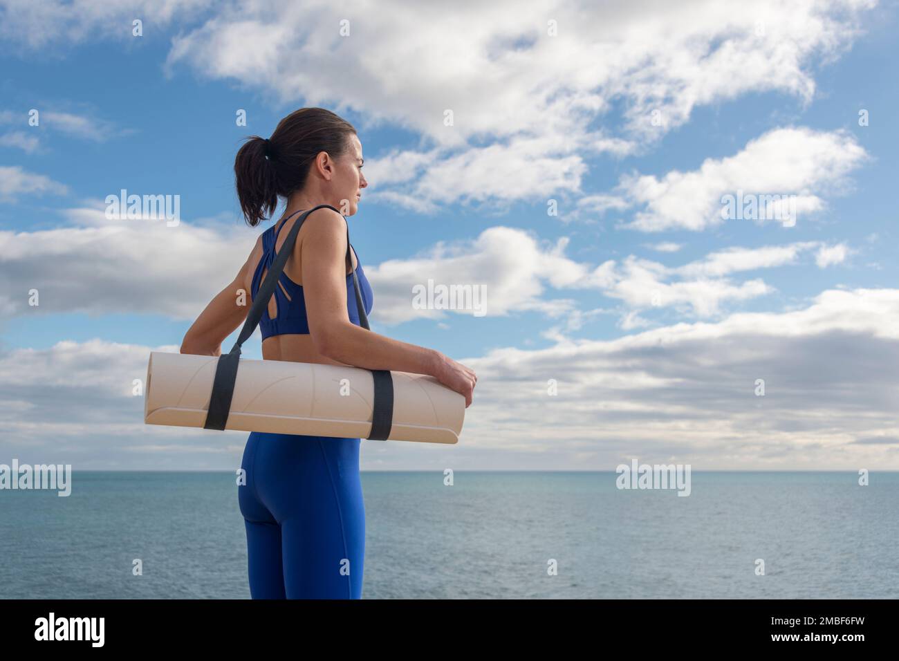 Donna in forma che trasporta un tappetino yoga che si prepara a praticare in riva al mare. Foto Stock