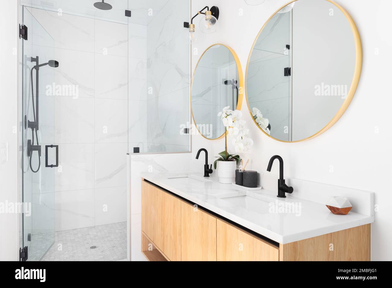 Una bella stanza da bagno con un mobile da toletta galleggiante in legno, specchi d'oro circolari, un ripiano in marmo e uno specchio rivestito in marmo con rubinetti neri. Foto Stock