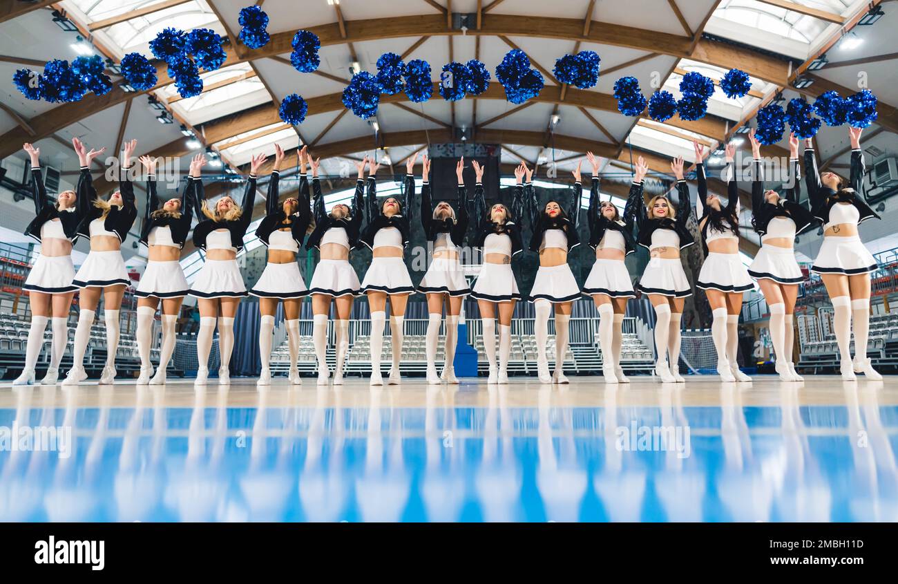 Vista frontale delle cheerleaders che lanciano i loro pom-poms blu in su nell'aria nella sala di sport. Primo piano sfocato. Foto di alta qualità Foto Stock