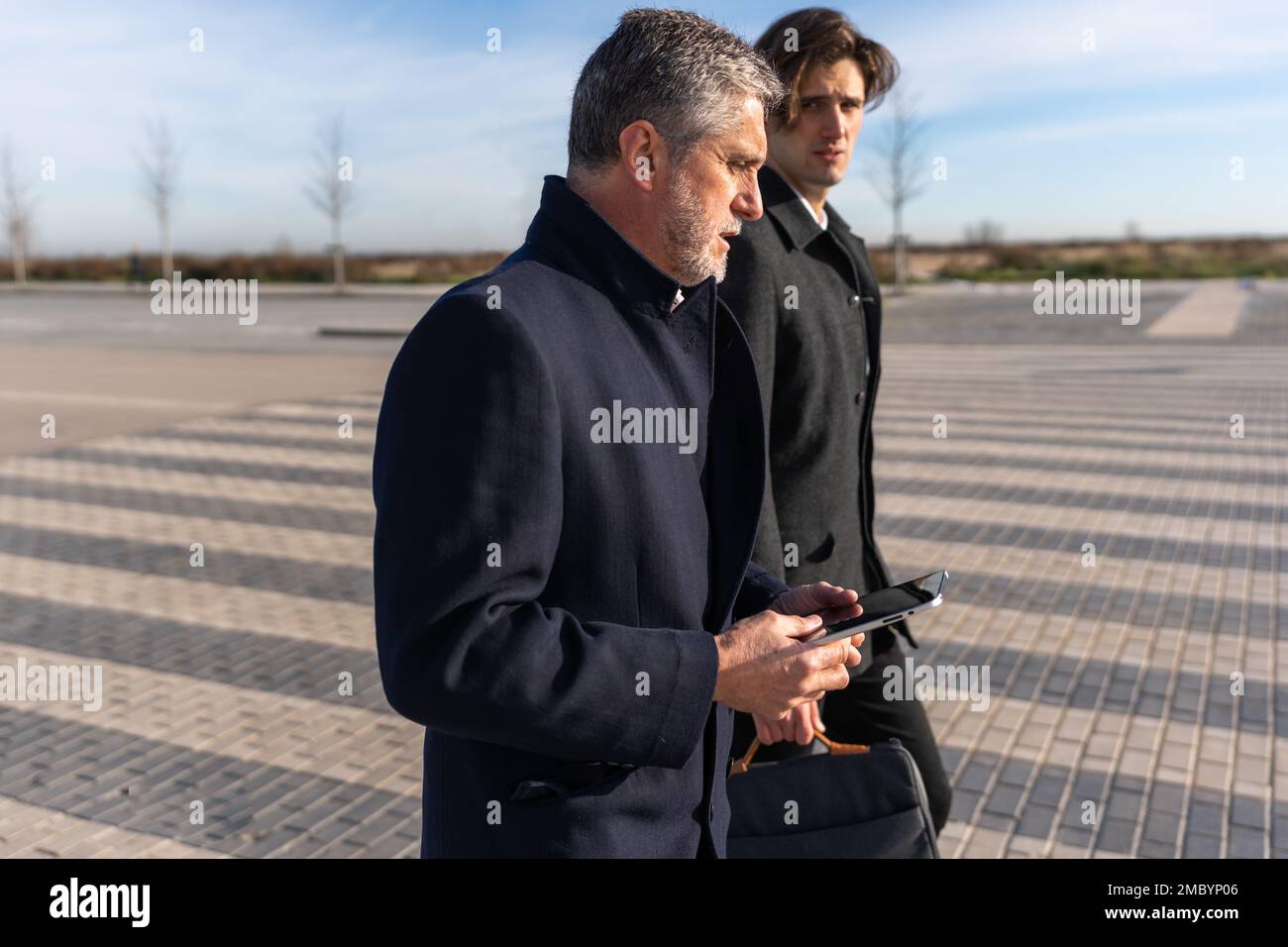 Uomo d'affari maturo e fiducioso in un elegante tablet per la navigazione mentre si cammina con un collega su strada asfaltata Foto Stock
