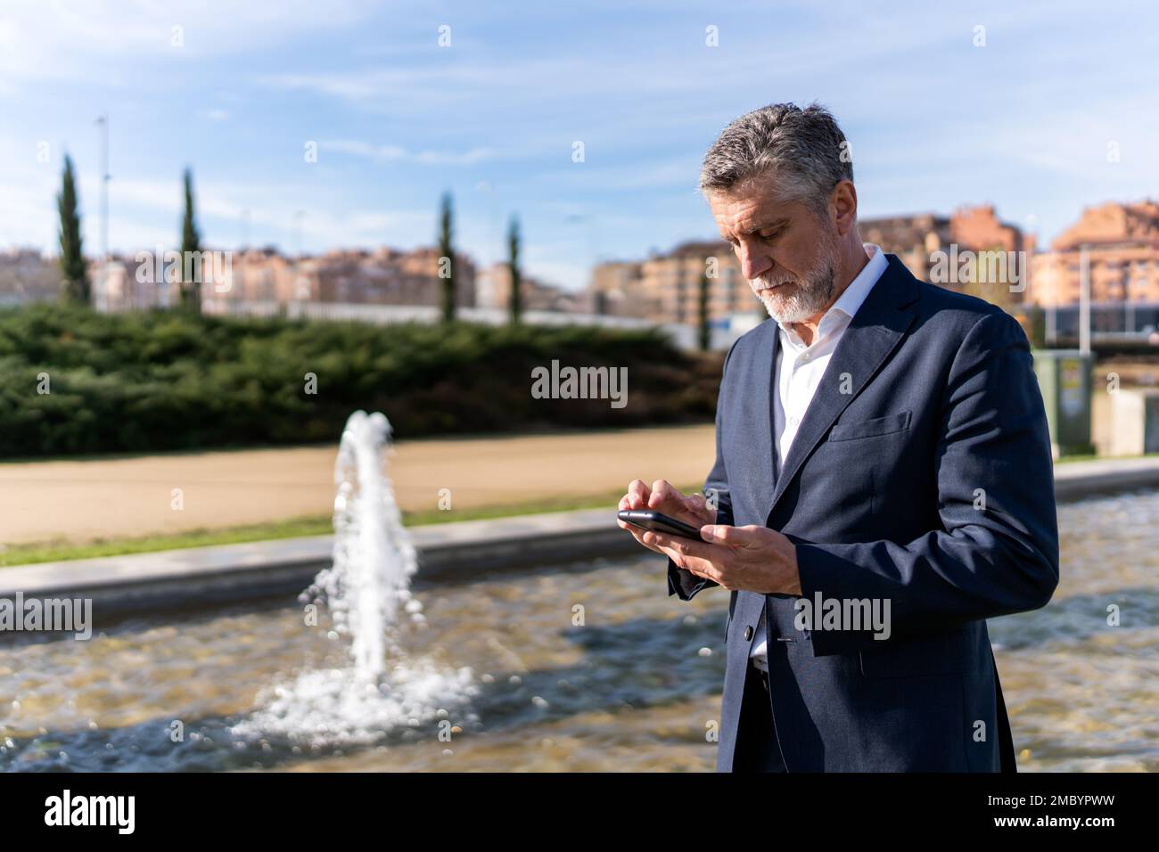 Serio uomo d'affari maturo in abbigliamento formale navigazione smartphone mentre in piedi sulla strada vicino fontana Foto Stock