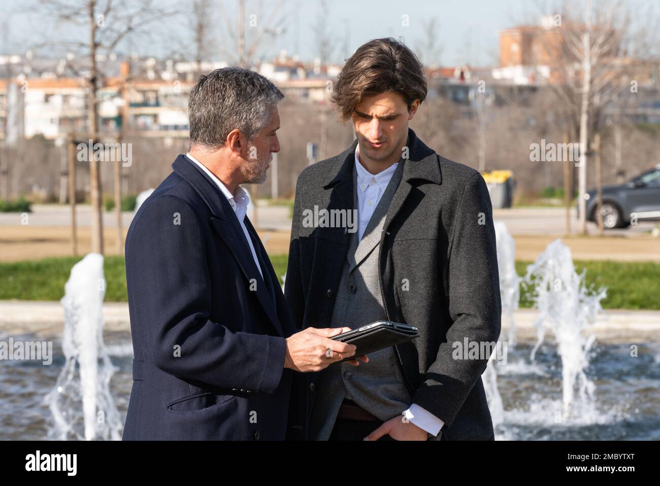 Uomini d'affari sicuri di indossare abiti formali in piedi sulla strada vicino alla fontana e controllare le informazioni sul tablet mentre si lavora in remoto dal parco Foto Stock