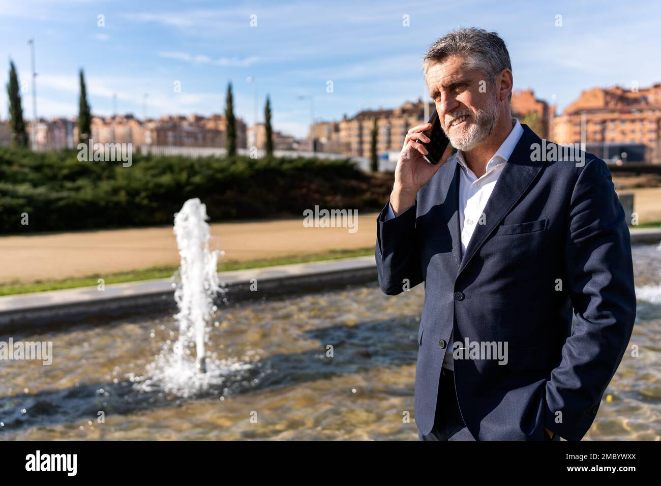 Uomo d'affari maturo allegro e bearded in abbigliamento formale sorridendo e guardando via mentre si parla sullo smartphone durante la passeggiata vicino alla fontana Foto Stock