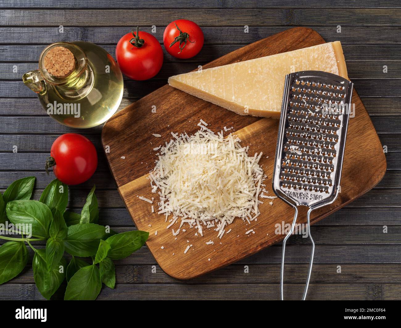 Parmigiano e grattugia su un tagliere. Formaggio grana padano intero e grattugiato, grattugia, olio d'oliva, basilico verde e pomodori su fondo ligneo. Foto Stock