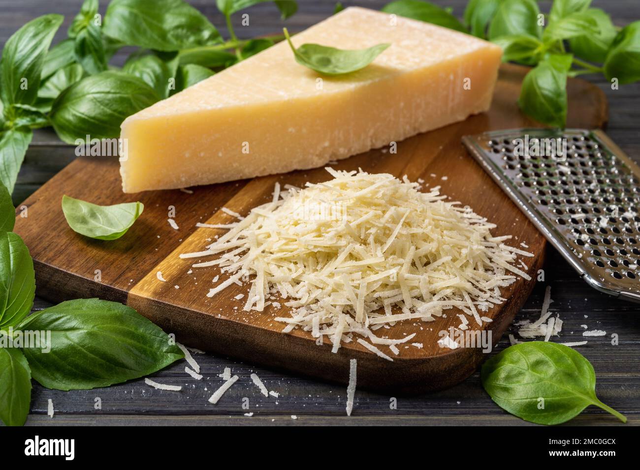 Parmigiano grattugiato e grattugia su un tagliere. Grattugia di grana padano a zeppa intera e grattugiata, grattugia in acciaio inox e primo piano di basilico verde. Foto Stock