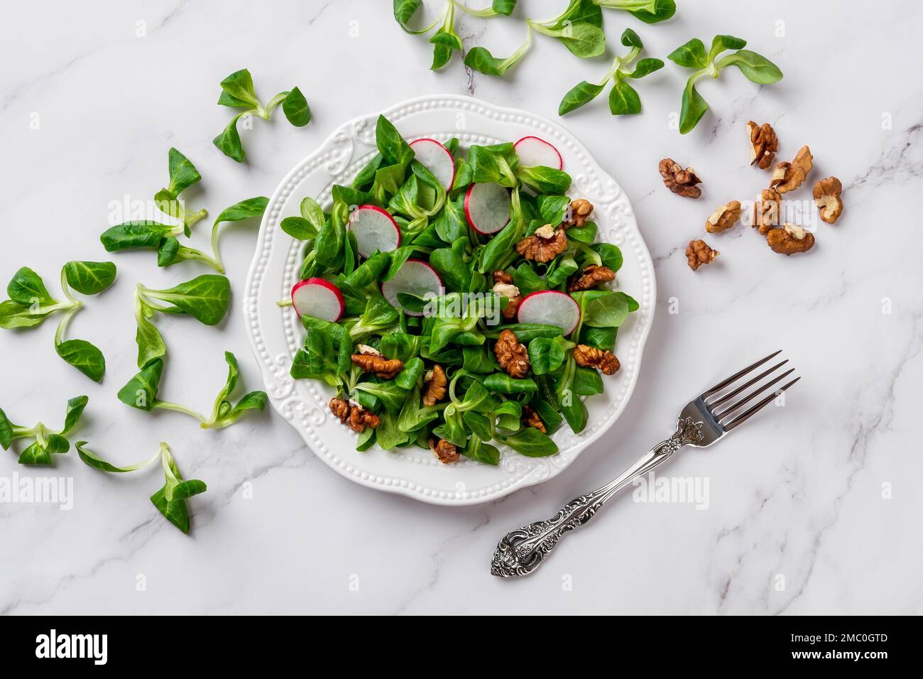 Insalata di lattuga di agnelli, ravanello e noci. Foglie di Corninsalata, rafano affettato e nocciole su un piatto bianco sopra la superficie di marmo. Ricetta di dieta dimagrante Foto Stock