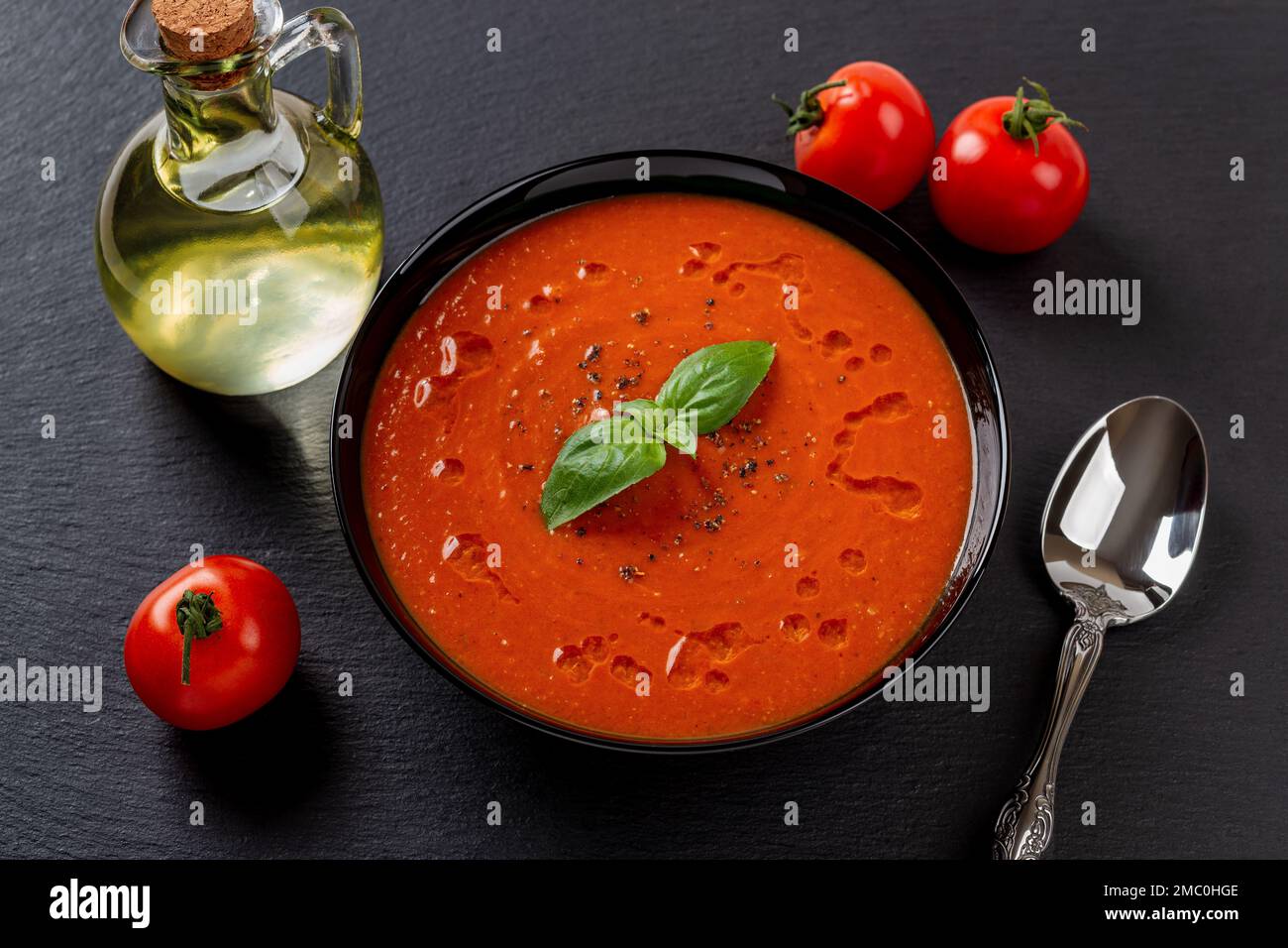 Zuppa di pomodoro calda e cucchiaio su sfondo nero. Porzione di purea di pomodori arrostiti, aglio e basilico appena fatto in un recipiente nero. Cibo vegano. Foto Stock