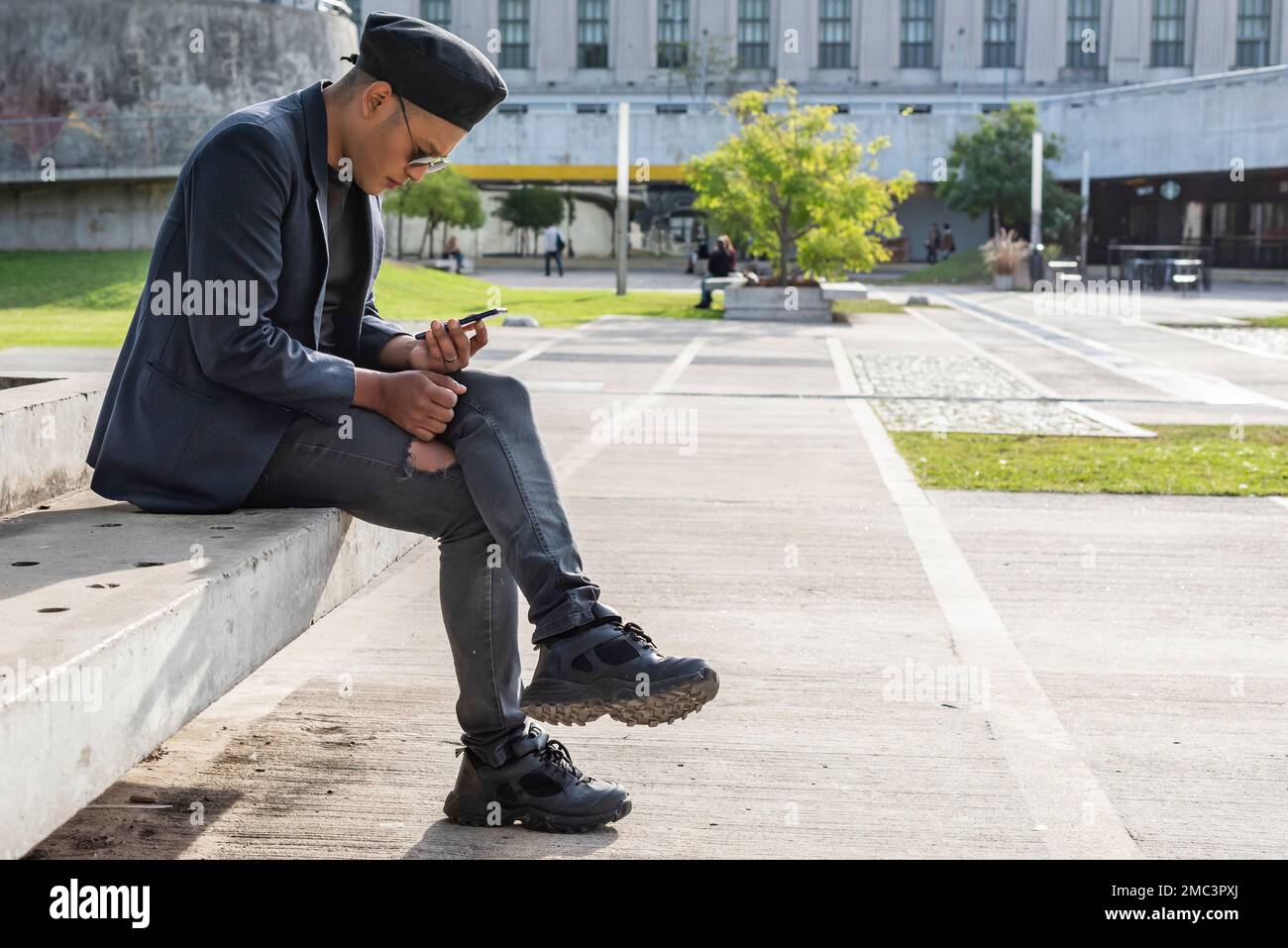 Giovane gay latino che indossa un cappello alla moda e occhiali da sole, che gli SMS sullo smartphone mentre si siede su una spiaggia all'aperto. LGTB. Foto Stock