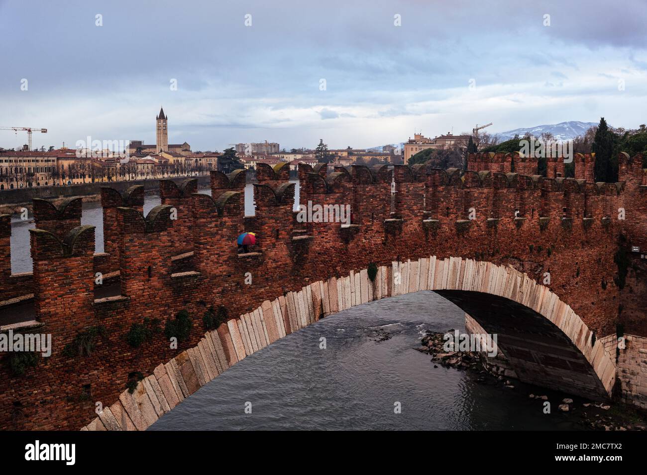 Verona, gennaio 2023. Una figura solitaria sotto un ombrello attraversa il ponte Scaligero sull'Adige in un suggestivo paesaggio urbano. Foto Stock