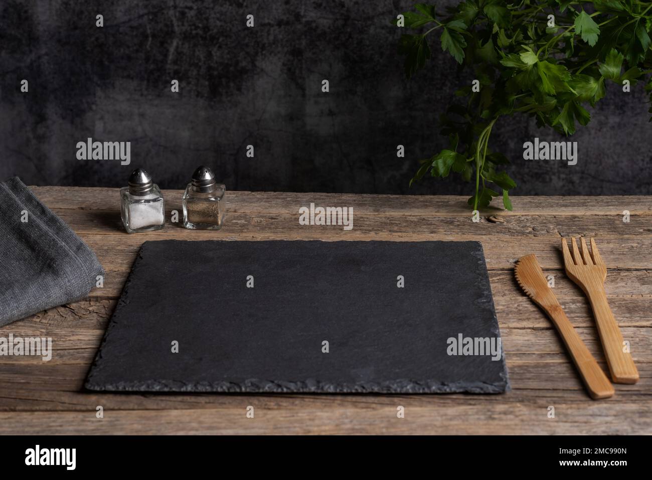 Piatto di pietra nera vuoto con posate di legno su un vecchio tavolo di legno rustico, accanto a sale, pepe e prezzemolo foglie. Foto Stock