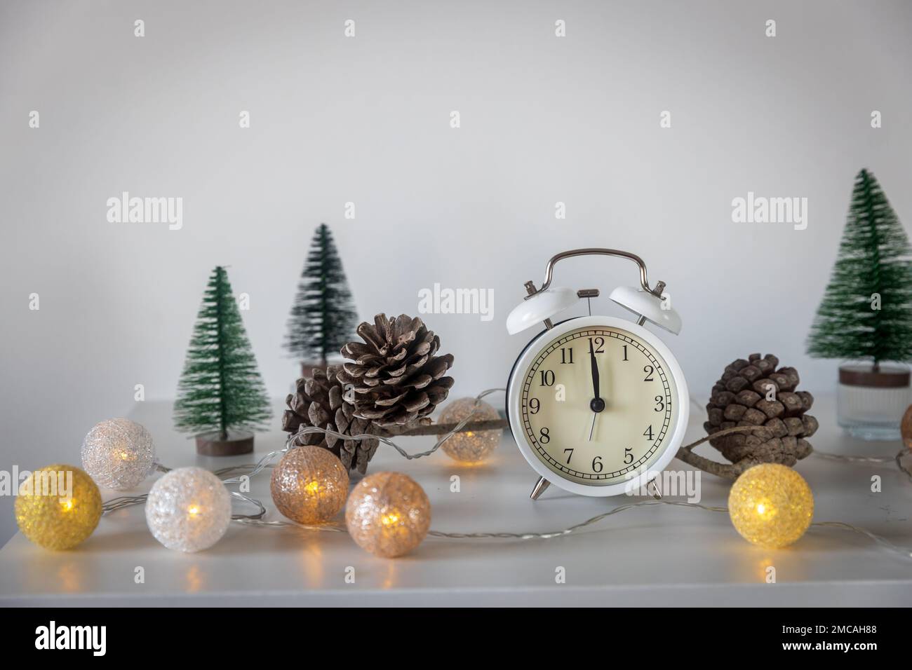 Una sveglia bianca, un puntatore a dodici, un piccolo albero di Natale verde artificiale e una ghirlanda su una cassettiera bianca. Spazio di copia. Biglietto di Natale. Foto Stock