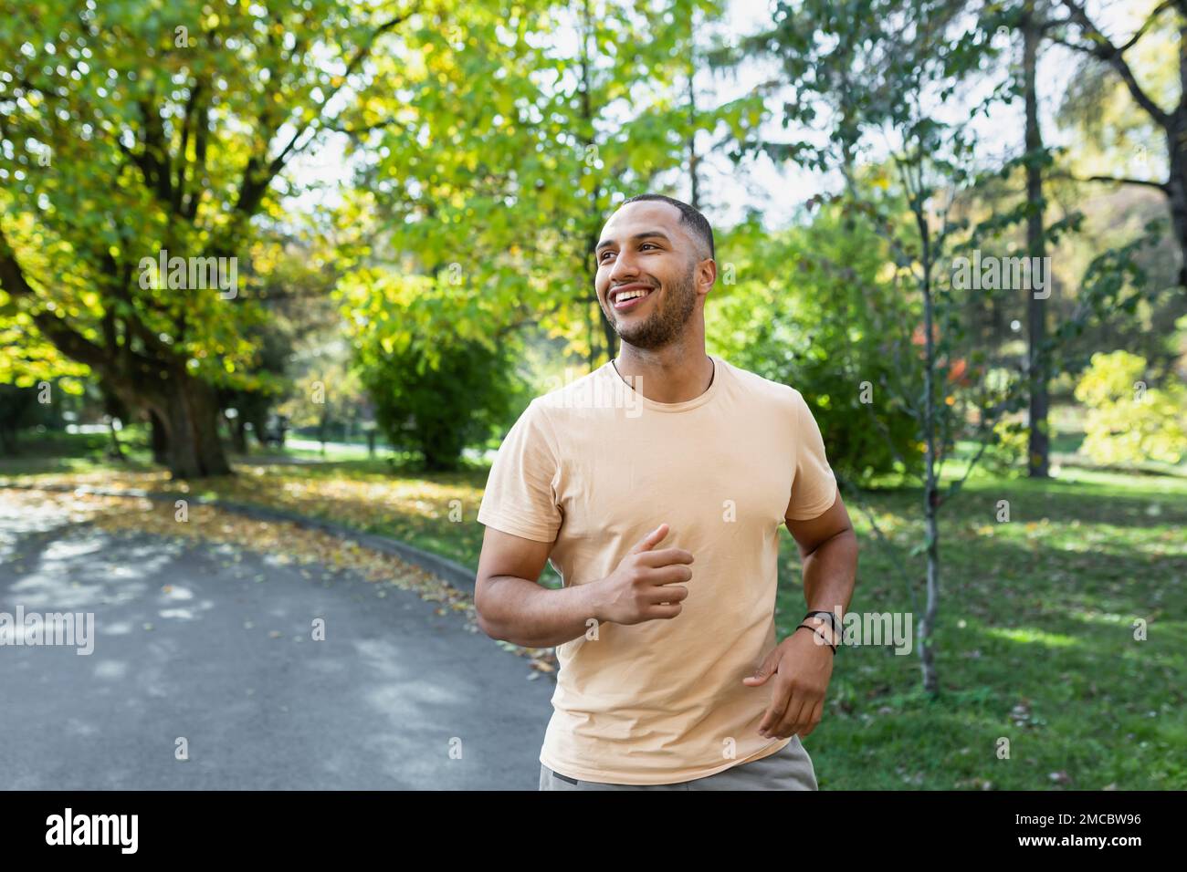 Allegro e di successo uomo ispanico jogging nel parco, uomo in esecuzione in una giornata di sole, sorridente e felice di avere un attività all'aperto. Foto Stock