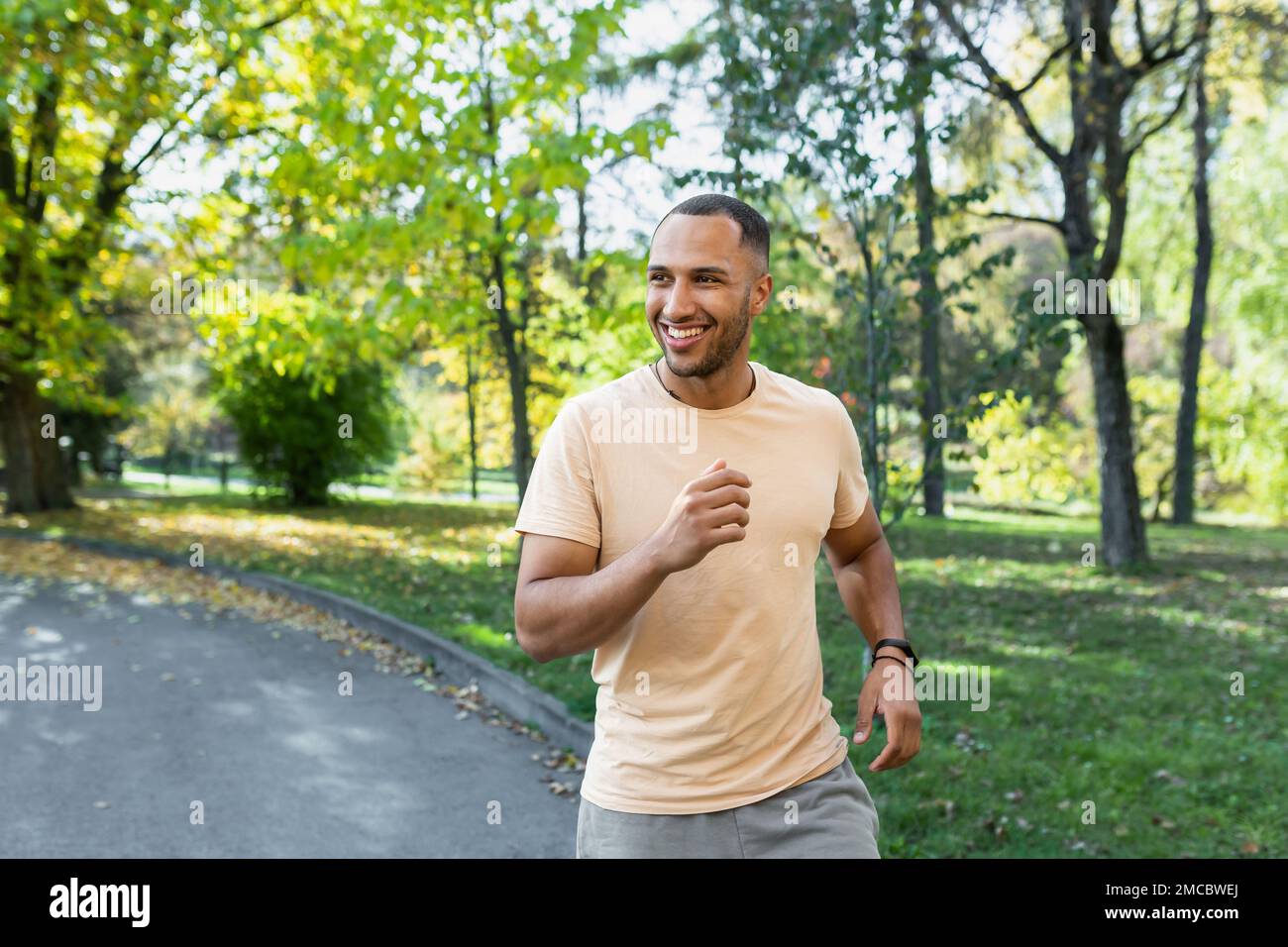 Allegro e di successo uomo ispanico jogging nel parco, uomo in esecuzione in una giornata di sole, sorridente e felice di avere un attività all'aperto. Foto Stock