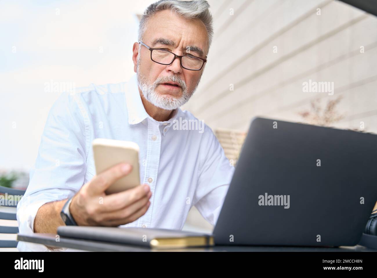 Uomo d'affari anziano che utilizza un telefono cellulare che lavora su un computer portatile all'aperto. Foto Stock