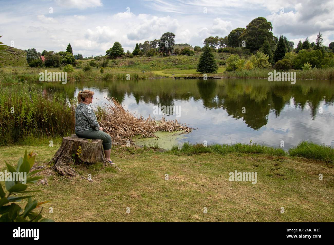 Dullstroom, Sud Africa - una donna matura siede su un ceppo e si affaccia su uno stagno Foto Stock