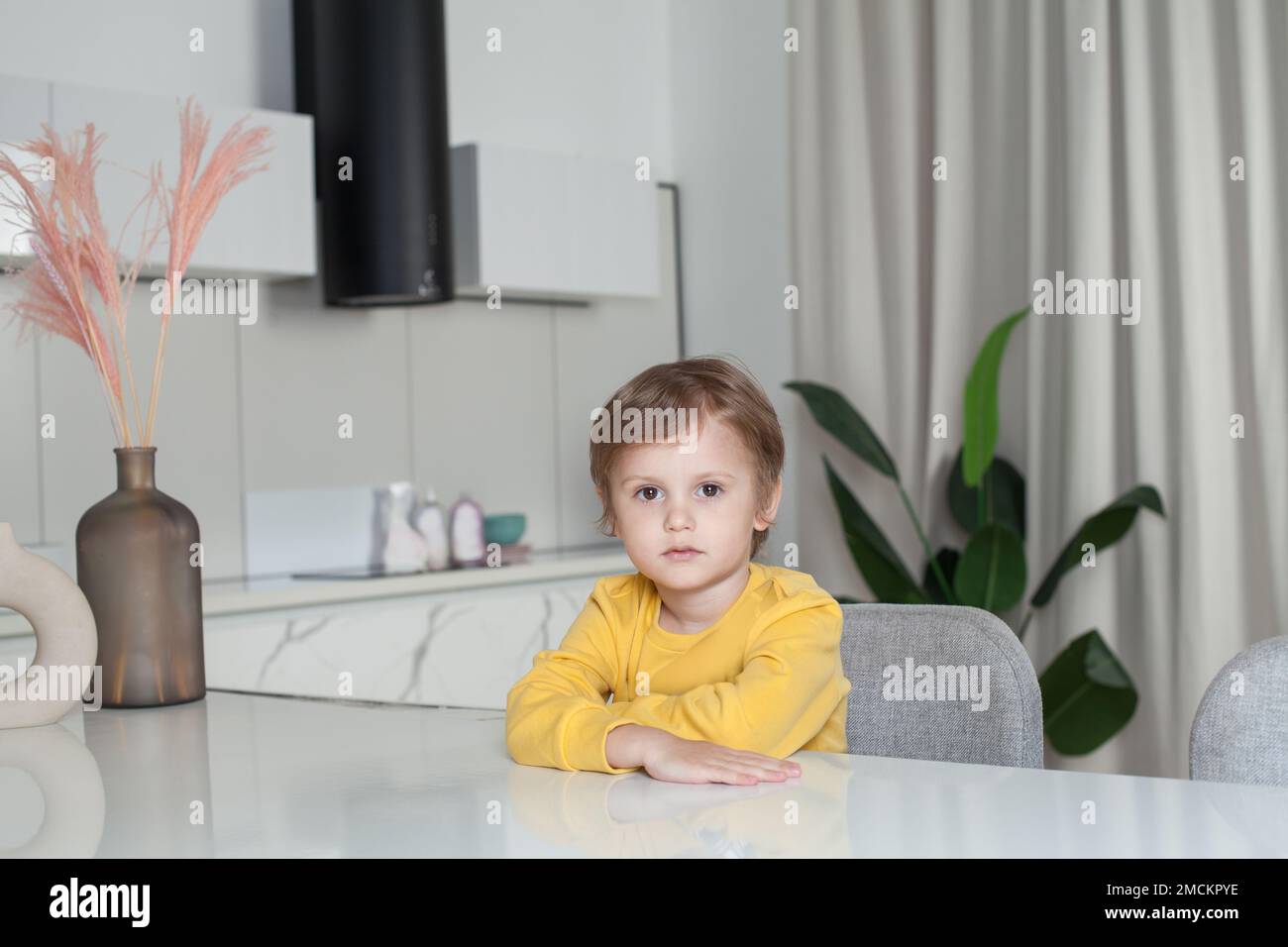 Bambino ragazzo pensivo con capelli castani in maglione giallo seduto accanto al tavolo su sfondo cucina studio Foto Stock