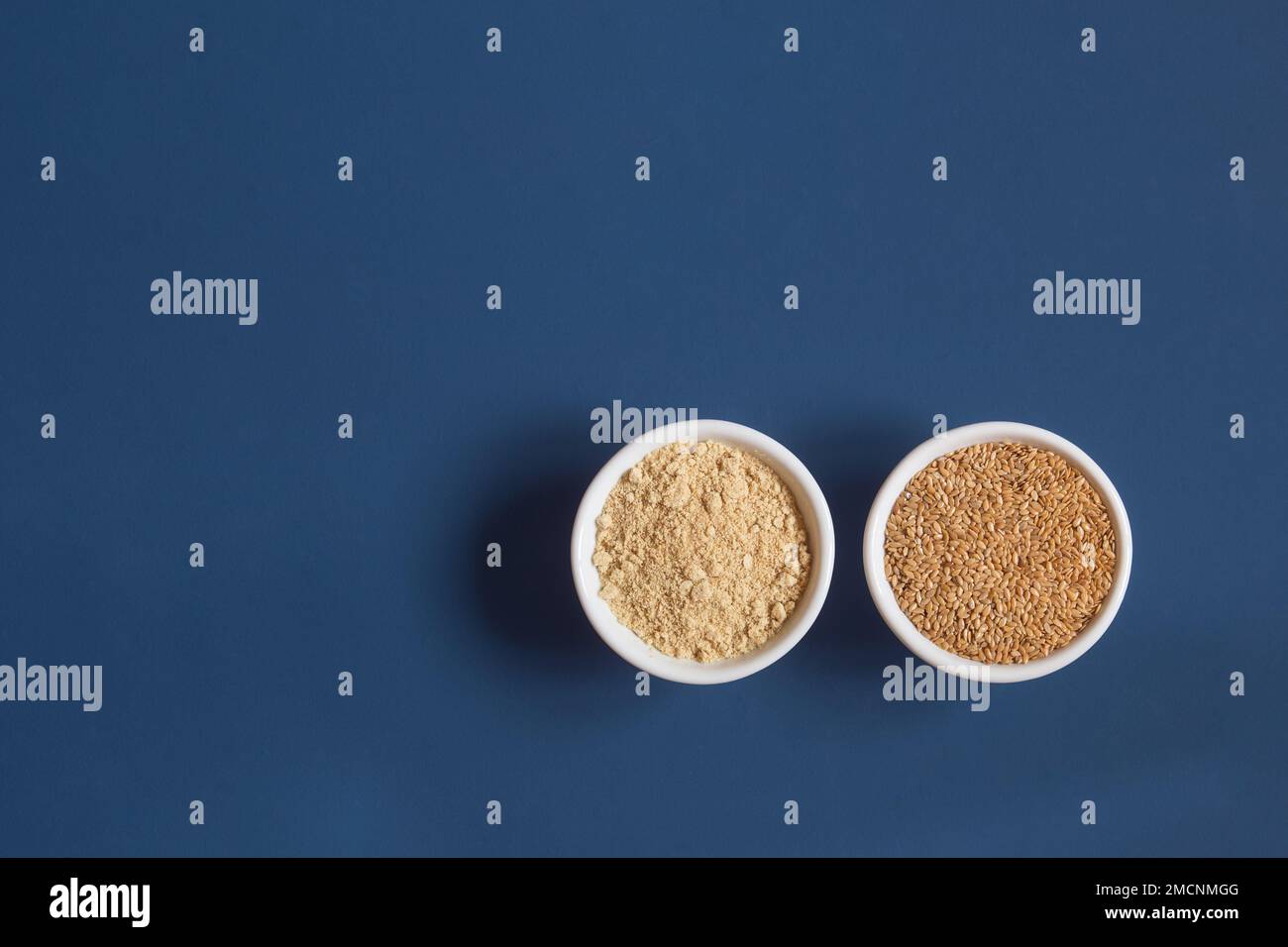 Vista dall'alto di un intero e schiacciato semi di lino su due diverse ciotole di ceramica cibo fianco a fianco su uno sfondo blu studio con spazio copia. Sano Foto Stock