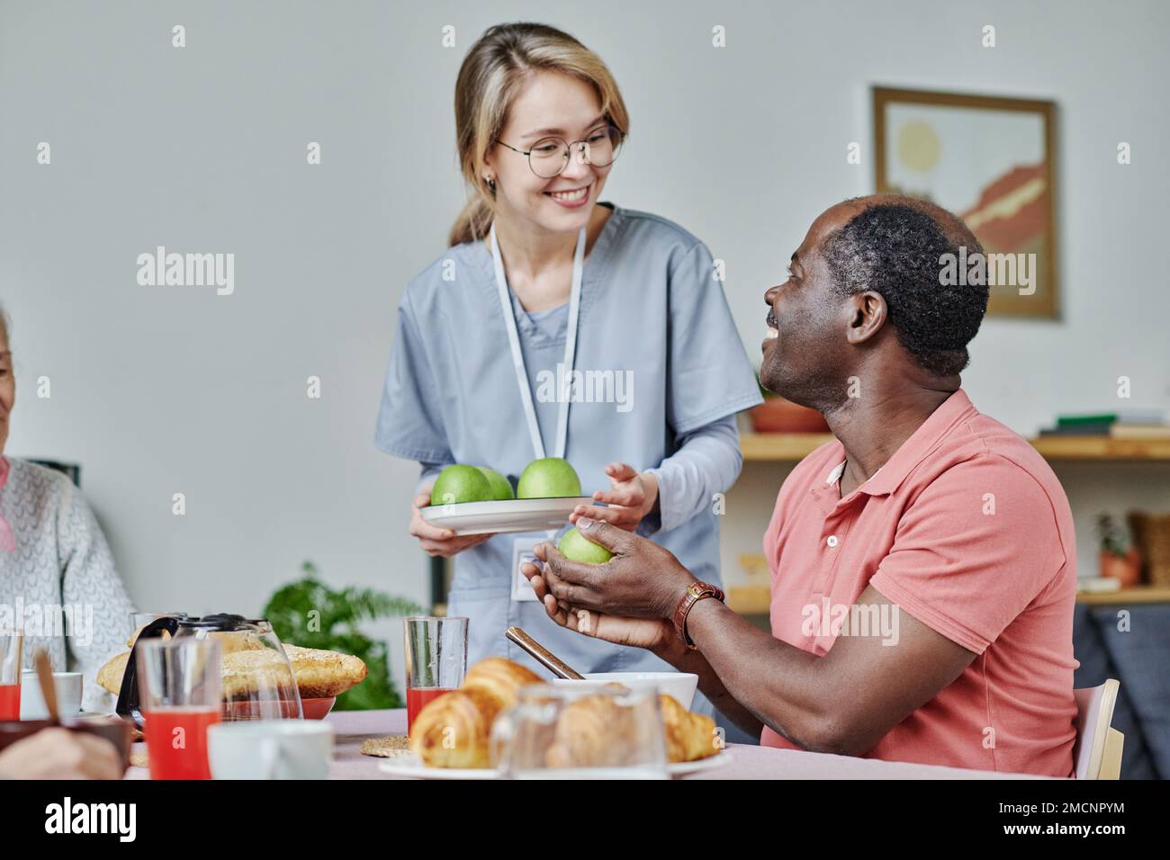 Sorridente caregiver che dà le mele all'uomo anziano mentre si siede al tavolo e mangia la sua colazione Foto Stock