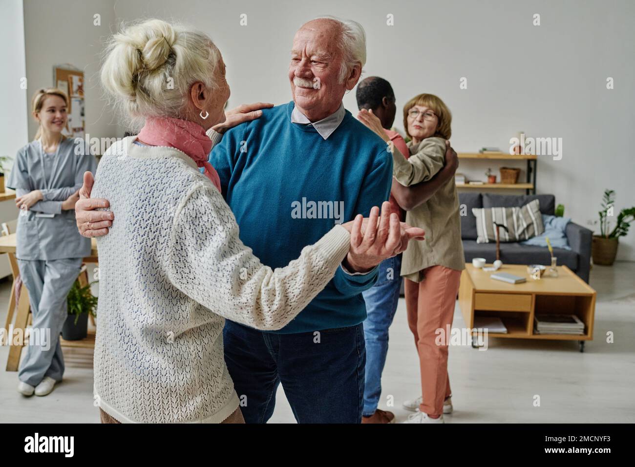 Felice coppia anziana ballare insieme in camera con altre persone e caregiver in background Foto Stock