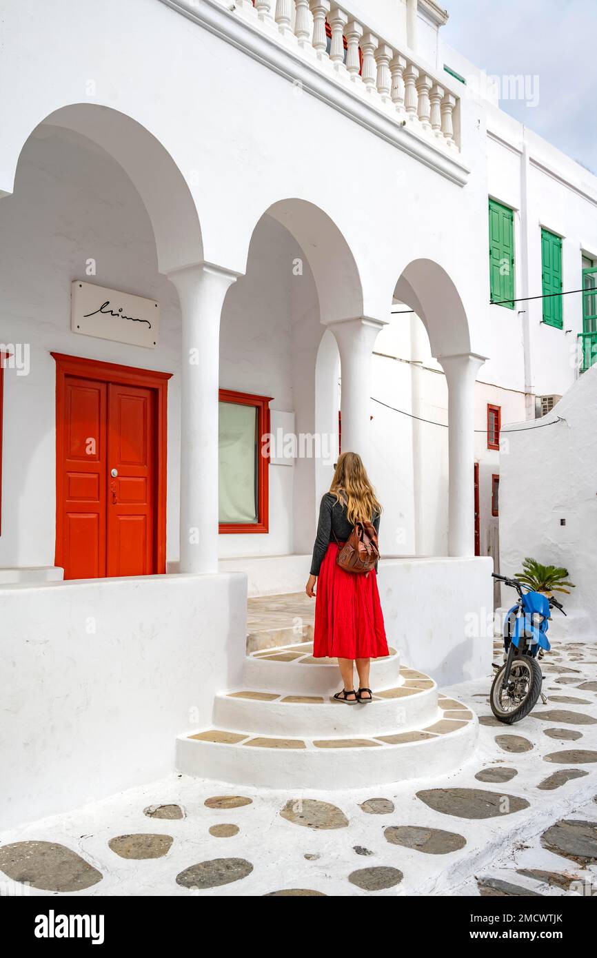 Giovane donna in gonna rossa di fronte a una casa cicladica bianca con colonne e porta rossa, vicoli della città vecchia Chora, Mykonos Town, Mykonos, Cicladi Foto Stock