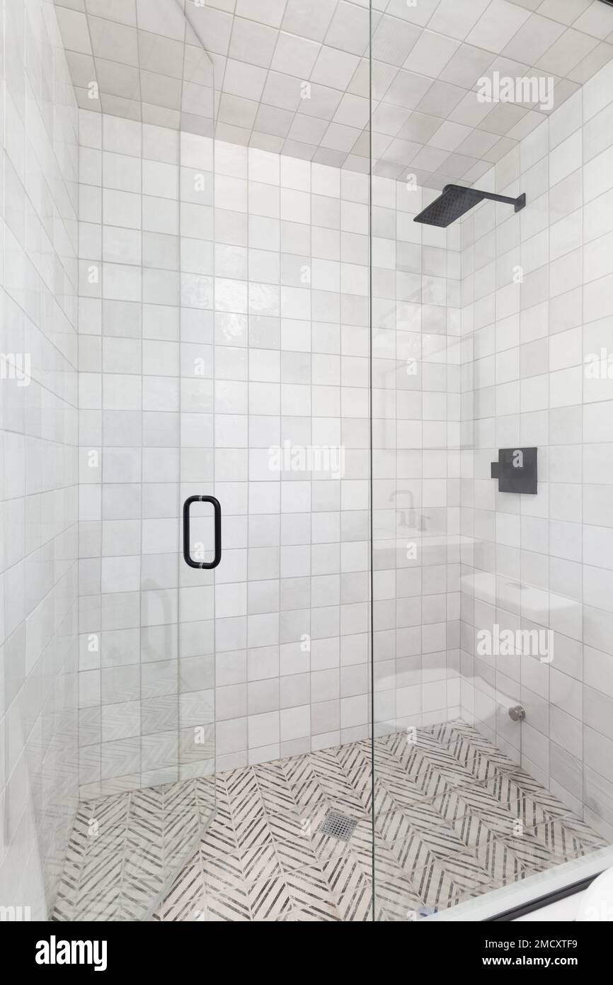 Una doccia con pareti piastrellate quadrate, pavimento piastrellato, rubinetto nero e porta in vetro. Foto Stock
