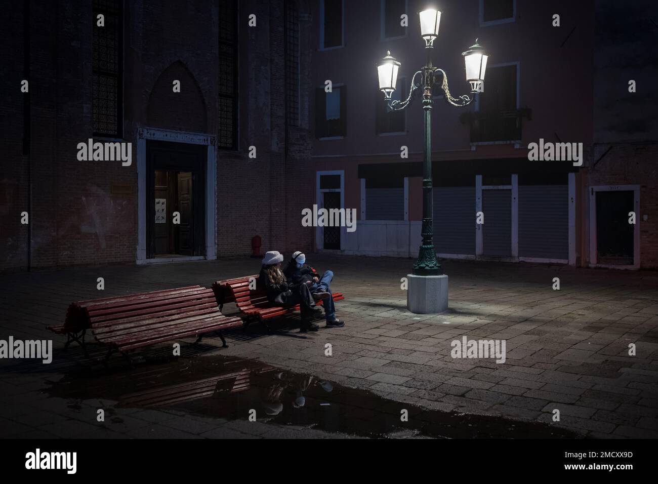 Gennaio 20, 2023. Un uomo e una donna siedono sotto una bella lampada da strada in una piazza deserta a Venezia, sotto un murale comunista e falce. Foto Stock