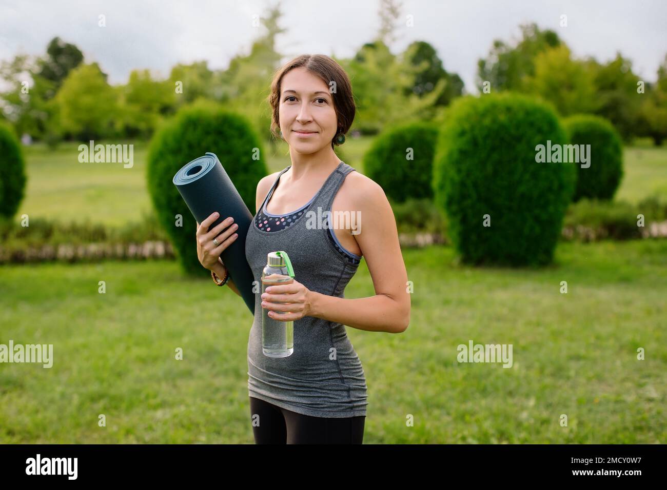 Una giovane ragazza atletica in una tuta grigia per il fitness sta andando a fare yoga in un parco verde, nelle sue mani una bottiglia d'acqua e un materassino yoga. Foto Stock