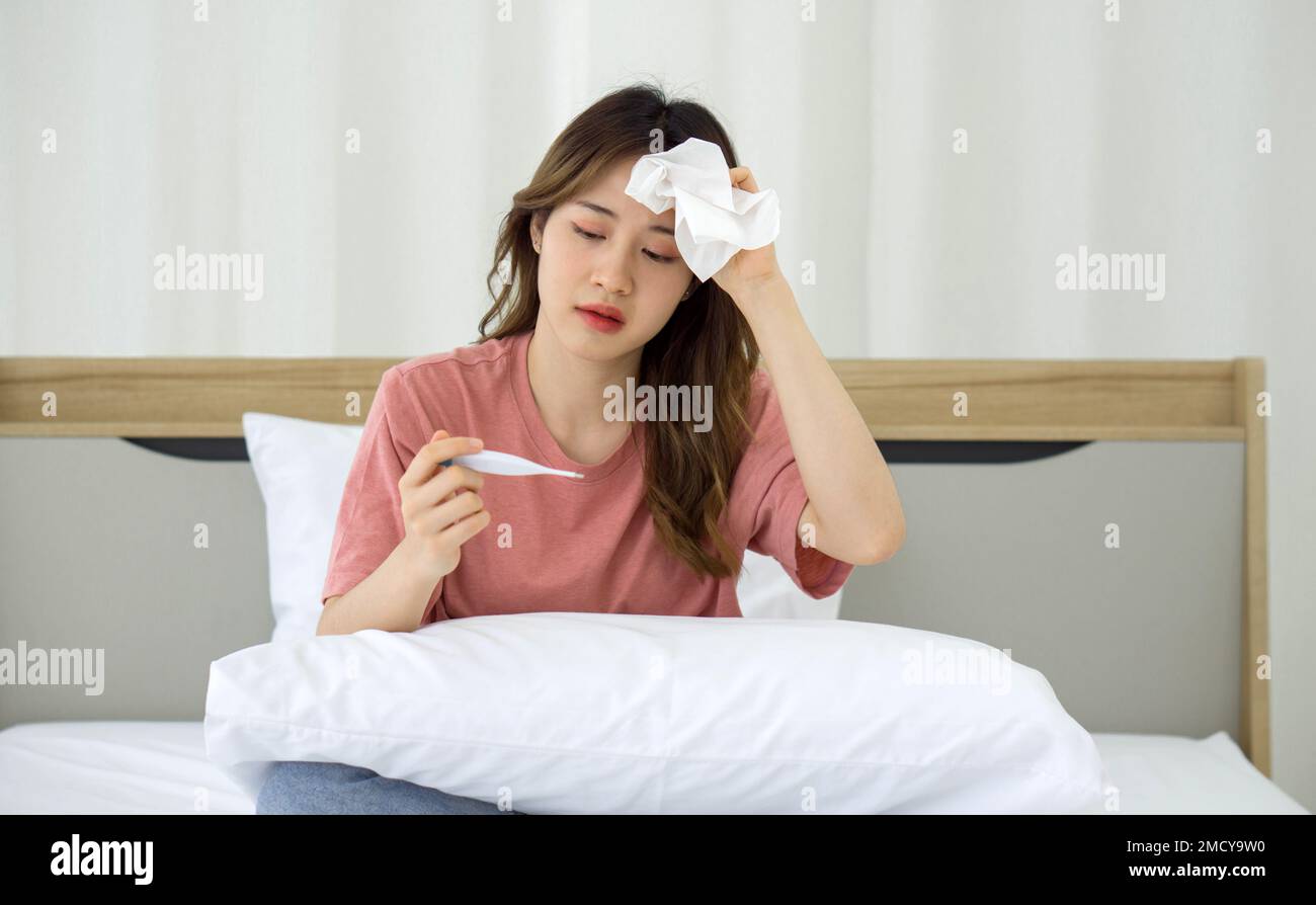 Giovane donna asiatica che prende la temperatura con un termometro digitale mentre si siede a letto. Covid-19 persone infette rimanere nelle loro camere da soli per la sicurezza, non Foto Stock