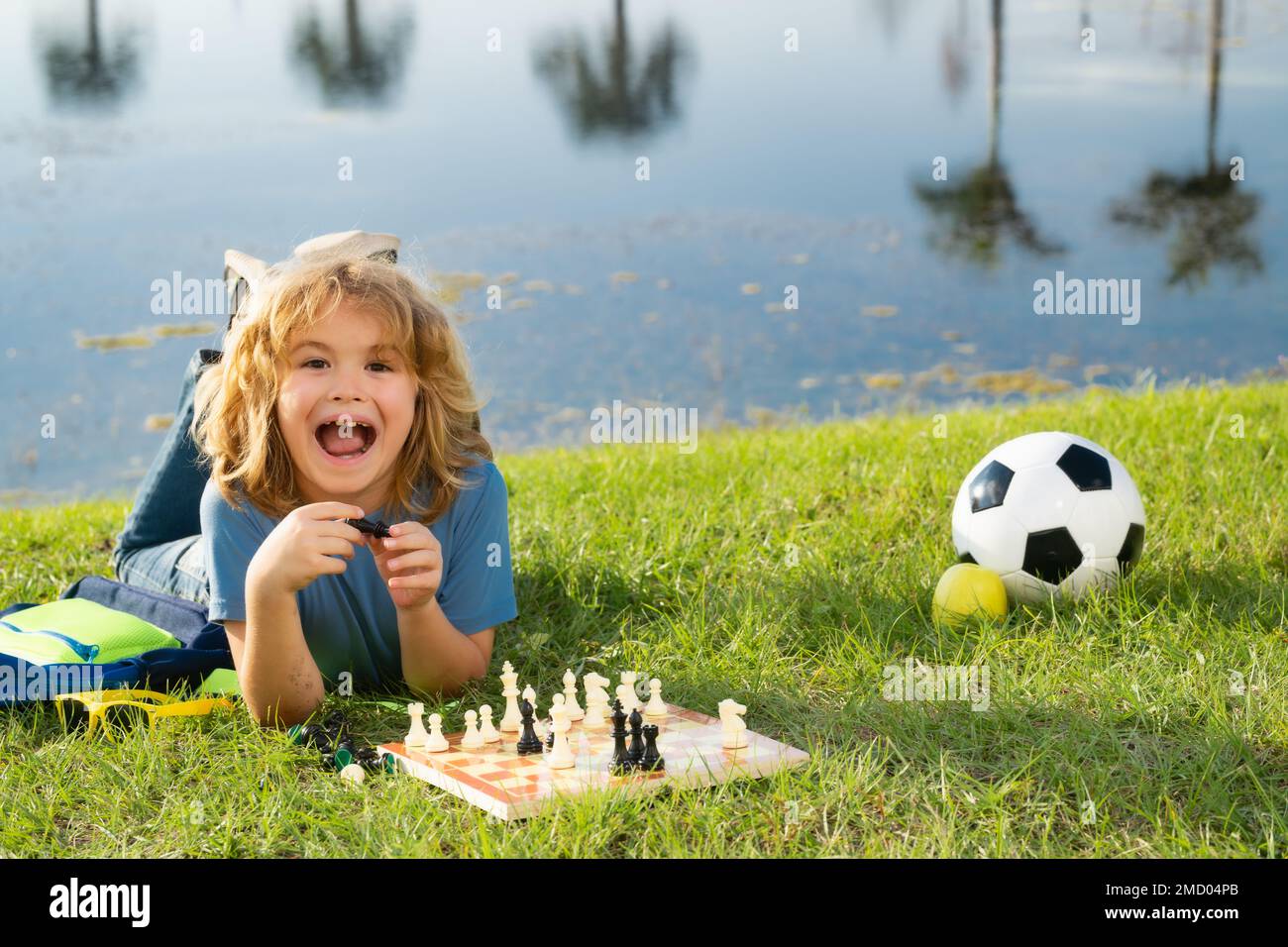 Ragazzo eccitato che gioca a scacchi nel cortile, adagiato sull'erba. Gioco di scacchi concentrato per bambini. Gioco da tavolo per bambini all'aperto. Foto Stock