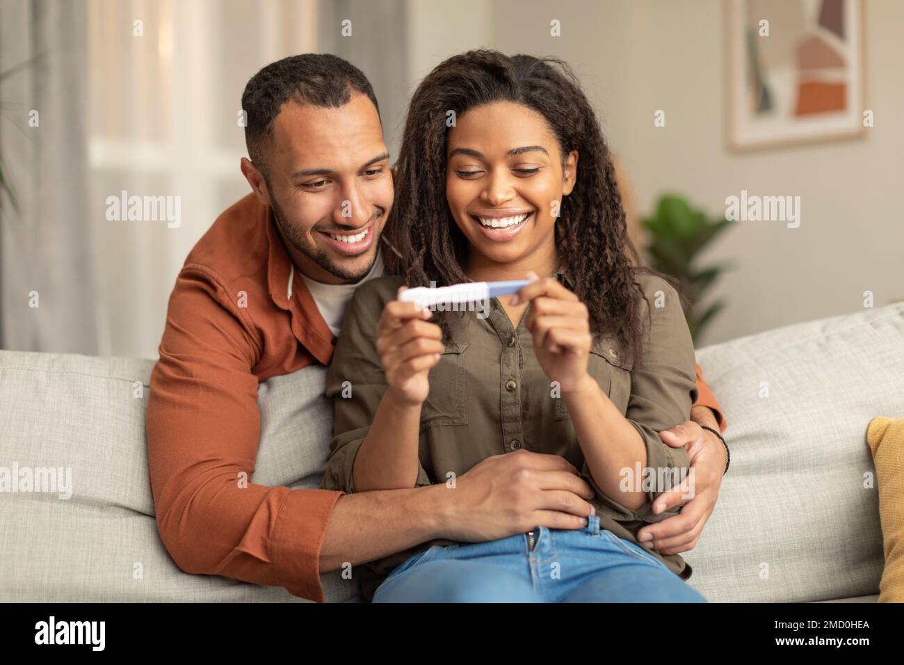 Felice coppia afro-americana che tiene test di gravidanza positivo e sorridente, uomo che abbraccia donna sul divano a casa Foto Stock