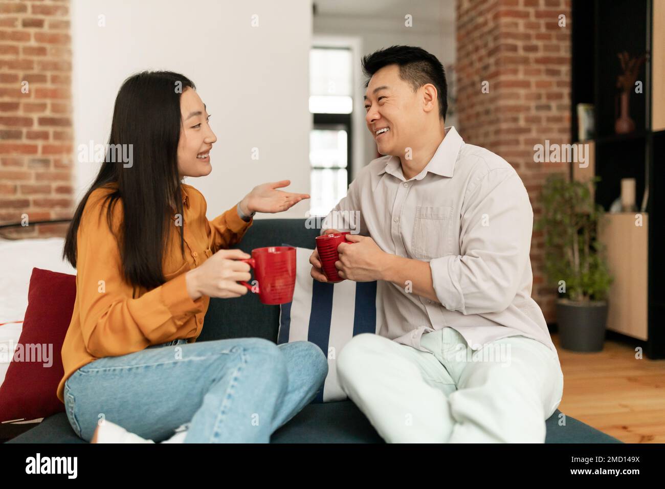 Sposi asiatici felici seduti sul divano con tazze di bevanda calda e di parlare, riposante nell'interno del soggiorno Foto Stock