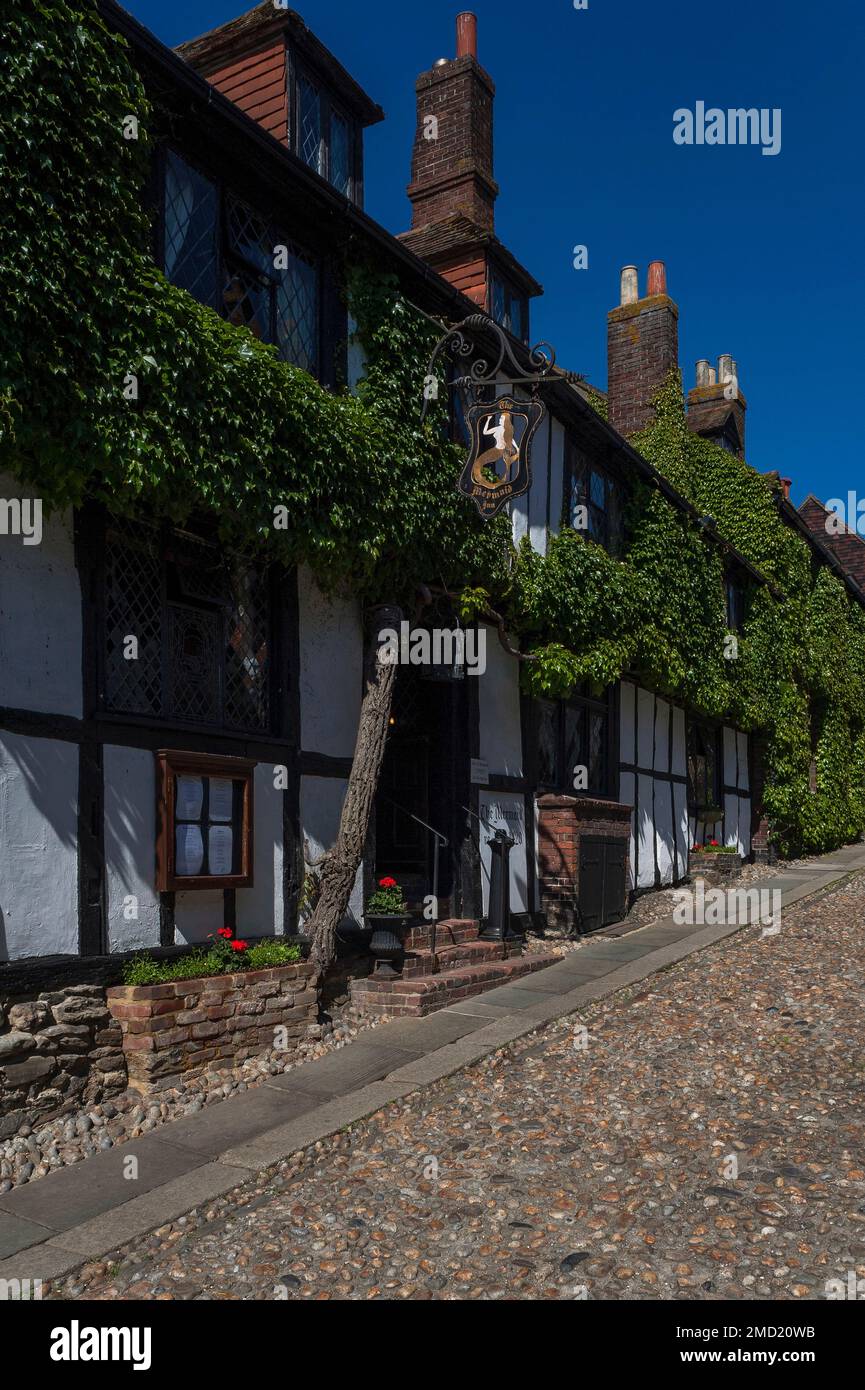 La glicine fiorisce attraverso la parte anteriore incorniciata in legno dell'antica Mermaid Inn in strada acciottolata Mermaid Street a Rye in East Sussex, Inghilterra, Regno Unito. L'insegna della locanda, che oscilla da elaborati lavori in ferro battuto, presenta una sirena dai capelli dorati con una coda dorata. Foto Stock