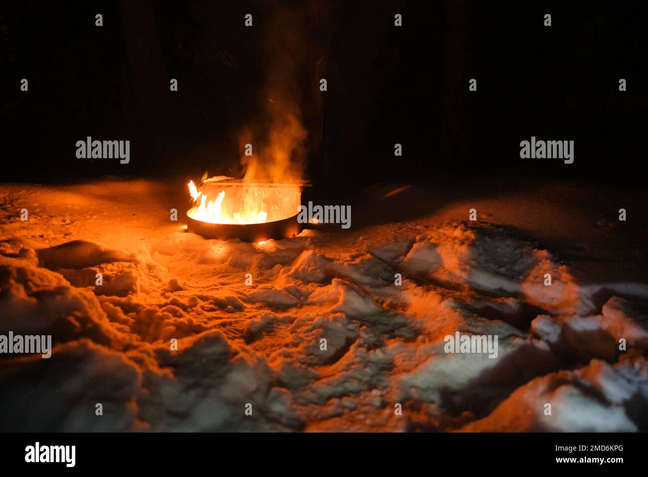 In inverno un incendio brucia in una fossa di un campeggio. La neve circonda il contenitore metallico dove le fiamme arancioni illuminano la neve circostante al buio. Foto Stock