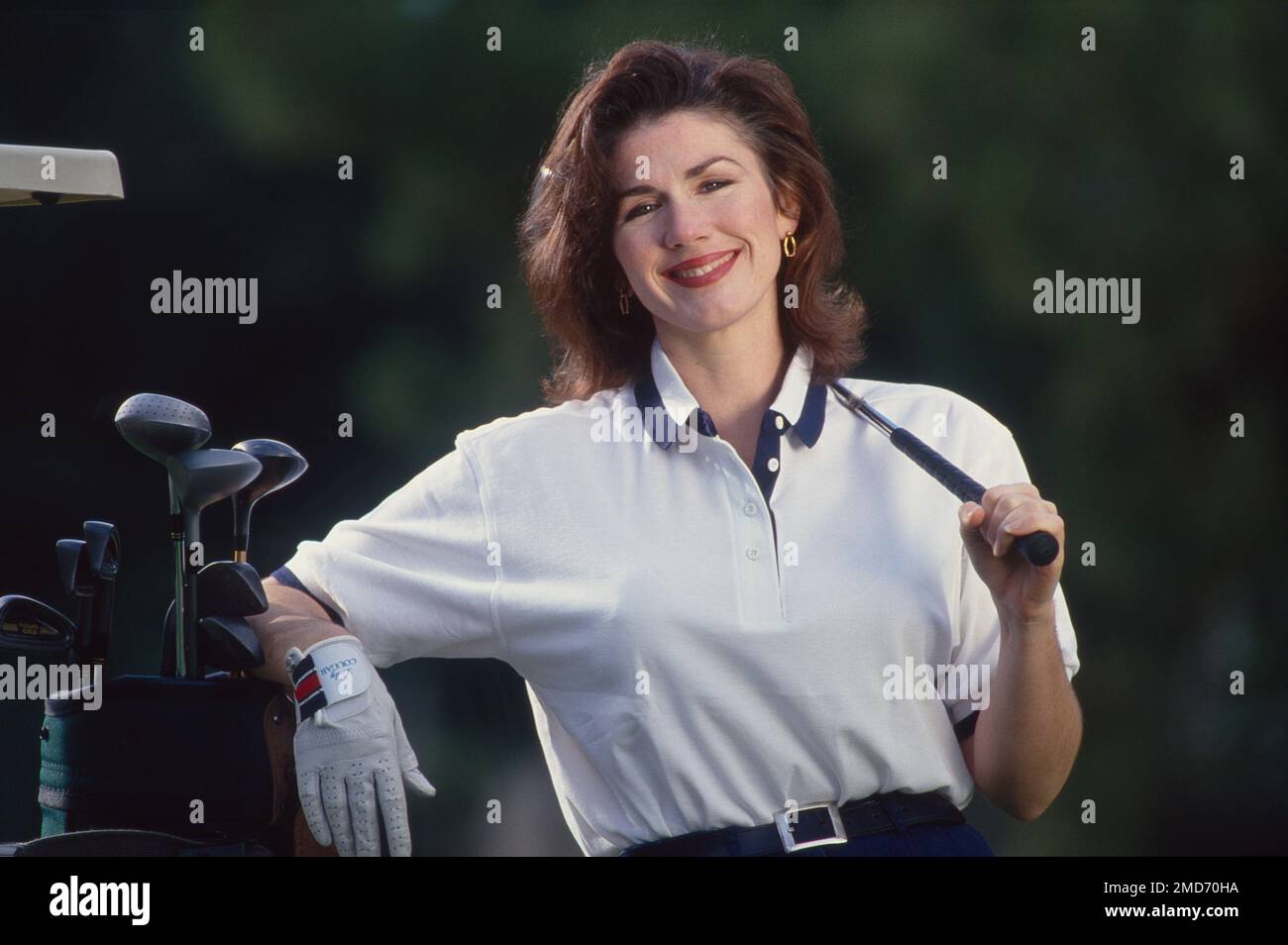 Donna giovane in posa mentre gioca a golf con il club sopra la sua spalla appoggiandosi su una borsa di club, sorridendo Foto Stock