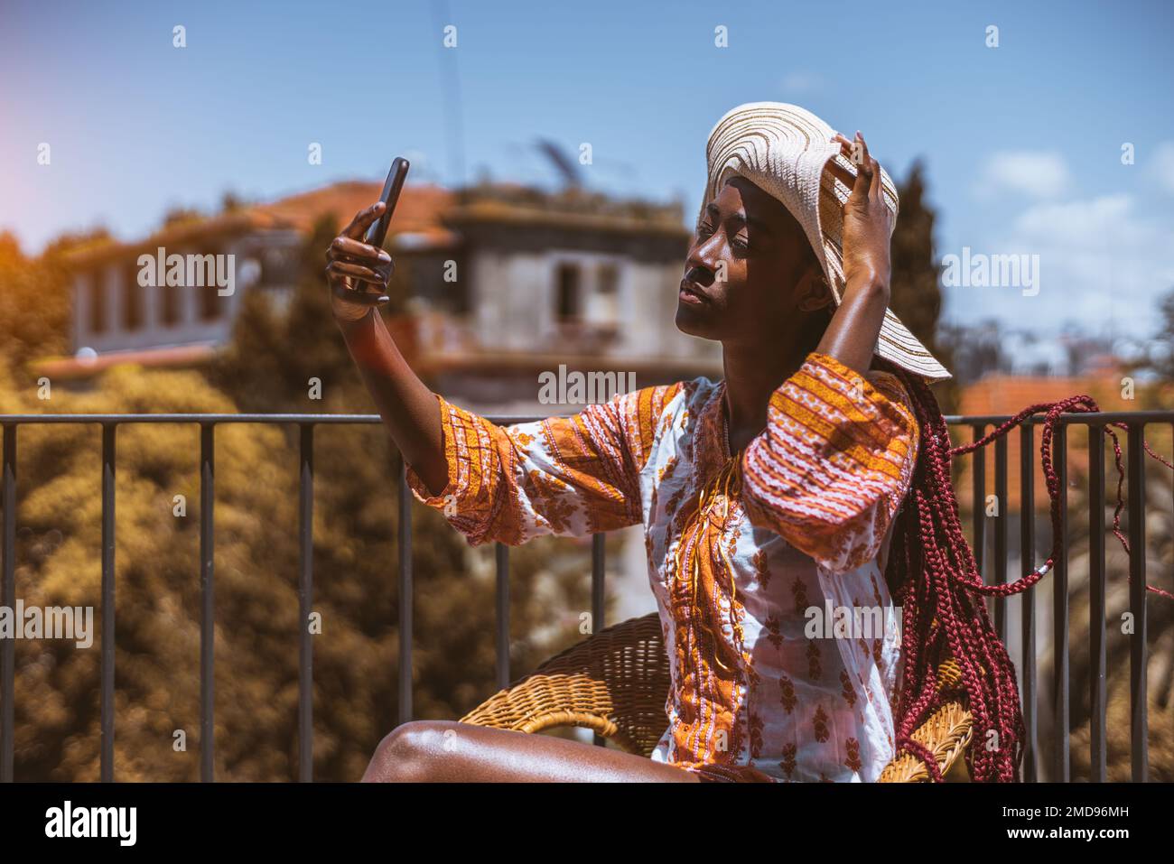 Una giovane donna, con lunghe trecce rosa, che indossa un abito corto colorato con maniche lunghe e un cappello largo-brimmed, che porta un selfie su una recinzione sul tetto Foto Stock