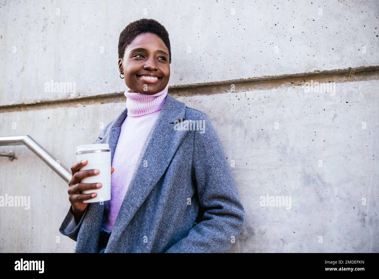 Felice afro americano femmina in cappotto elegante e maglione con zero rifiuti tazza di caffè sorridente e guardando via. Si appoggia su un muro di cemento sulla strada cittadina Foto Stock