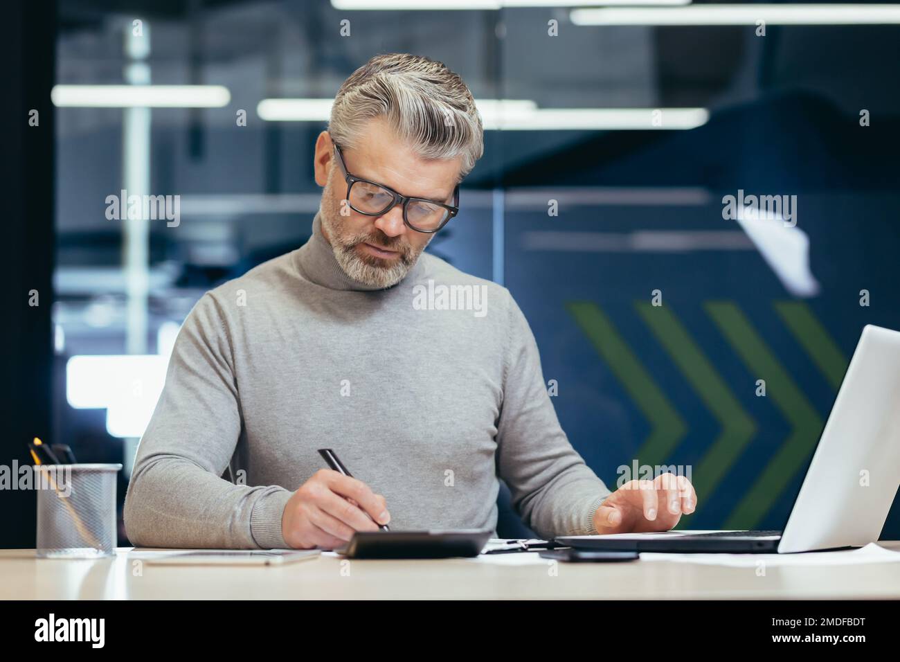 Uomo d'affari serio e pensante all'interno dell'ufficio al lavoro con un computer portatile, un uomo anziano dai capelli grigi che lavora seduto alla scrivania. Foto Stock