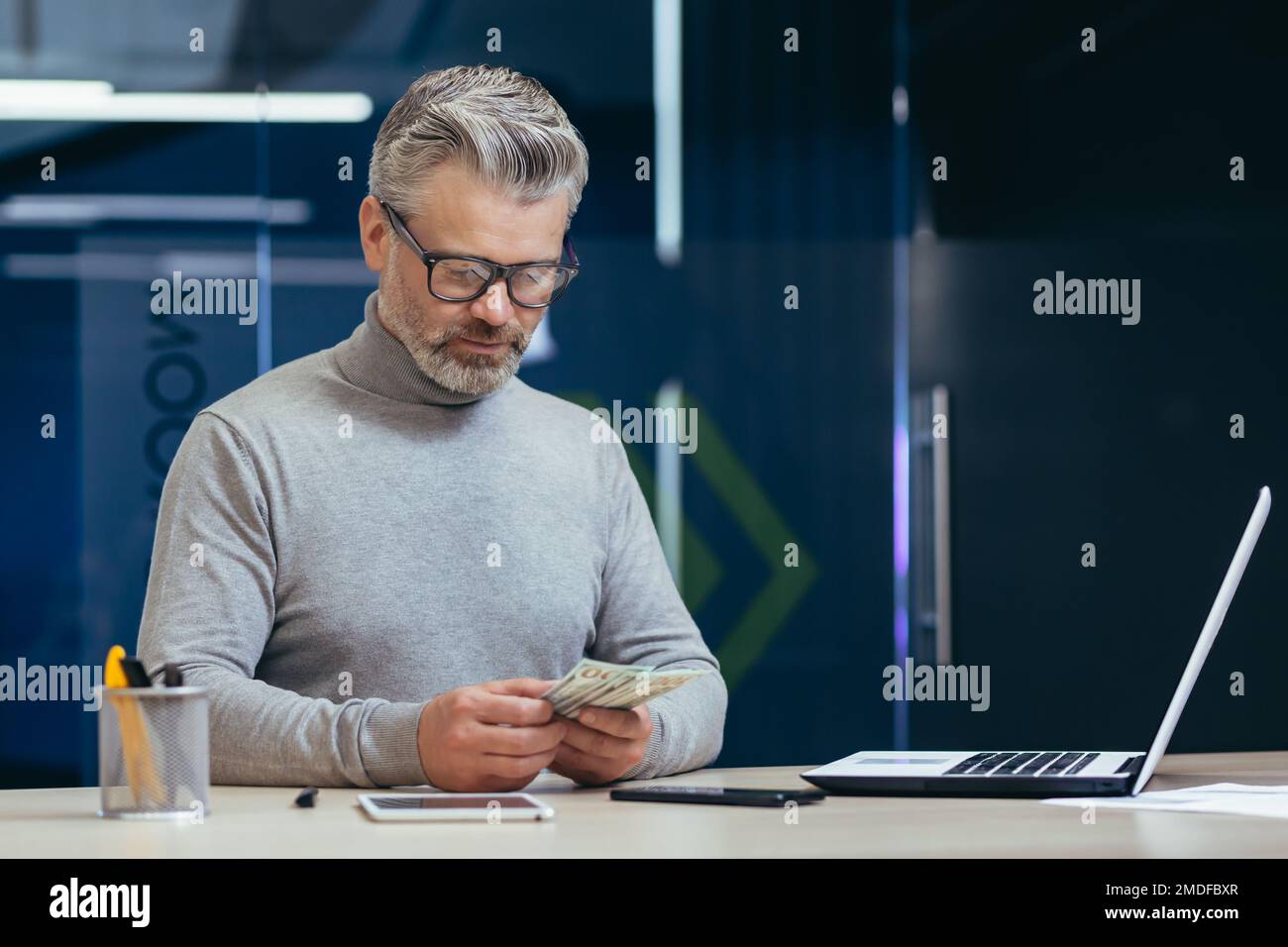 Investitore serio maturo che lavora all'interno dell'ufficio, uomo d'affari grigio alto che conta i dollari di contanti dei soldi, uomo d'affari che lavora all'interno dell'ufficio contemporaneo usando il laptop al lavoro. Foto Stock