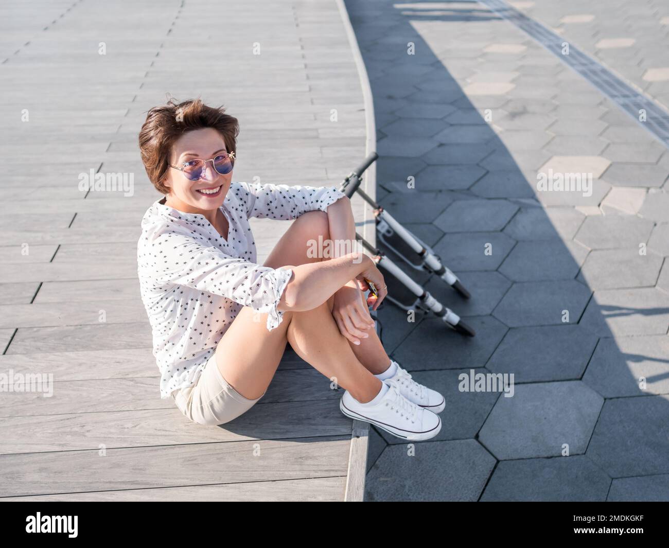 Vento ruffa capelli corti di donna in occhiali da sole colorati. Donna sorridente in una scena di legno aperto. Atmosfera estiva. Emozioni sincere. Calcio scooter - eco-amico Foto Stock