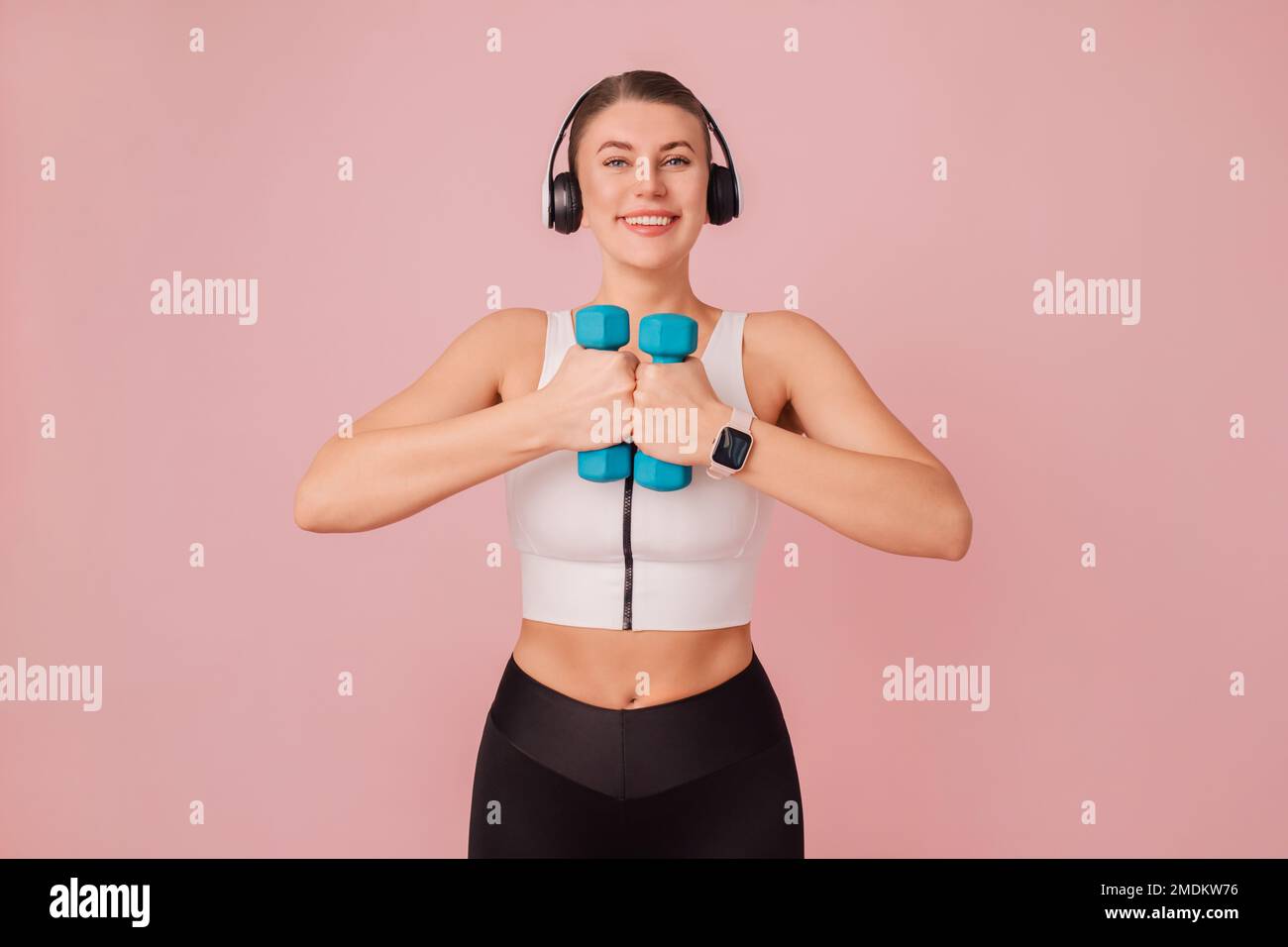 Ritratto di felice signora caucasica in abbigliamento sportivo con manubri blu su sfondo rosa. Giovane donna allegra con cuffie che indossano abbigliamento sportivo Foto Stock