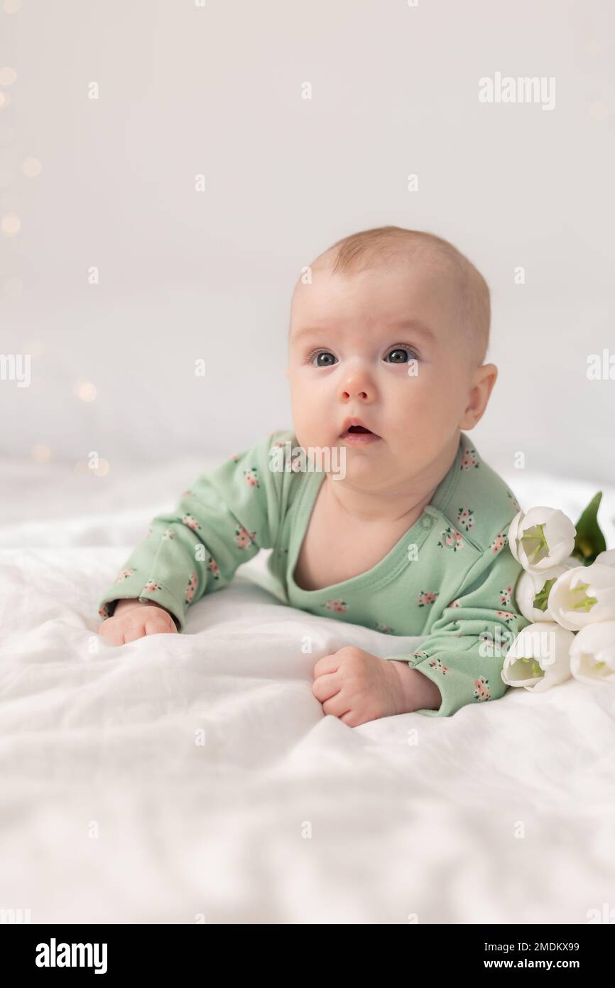 bambino carino in un bodysuit di cotone verde a casa su un letto bianco con tulipani. Primavera, Festa della mamma, 8 marzo. Foto di alta qualità Foto Stock