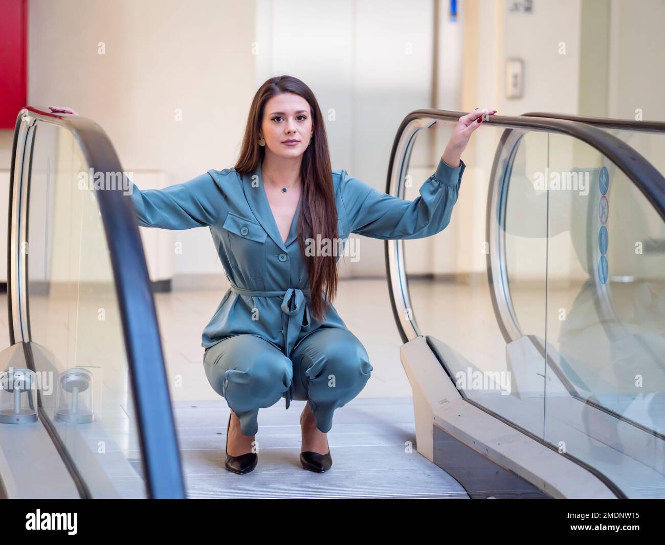 Giovane donna fantasiosa in negozio vuoto squatting squat spargere mani braccia prima scala mobile scala scale guardando la fotocamera occhio occhi contatto Foto Stock