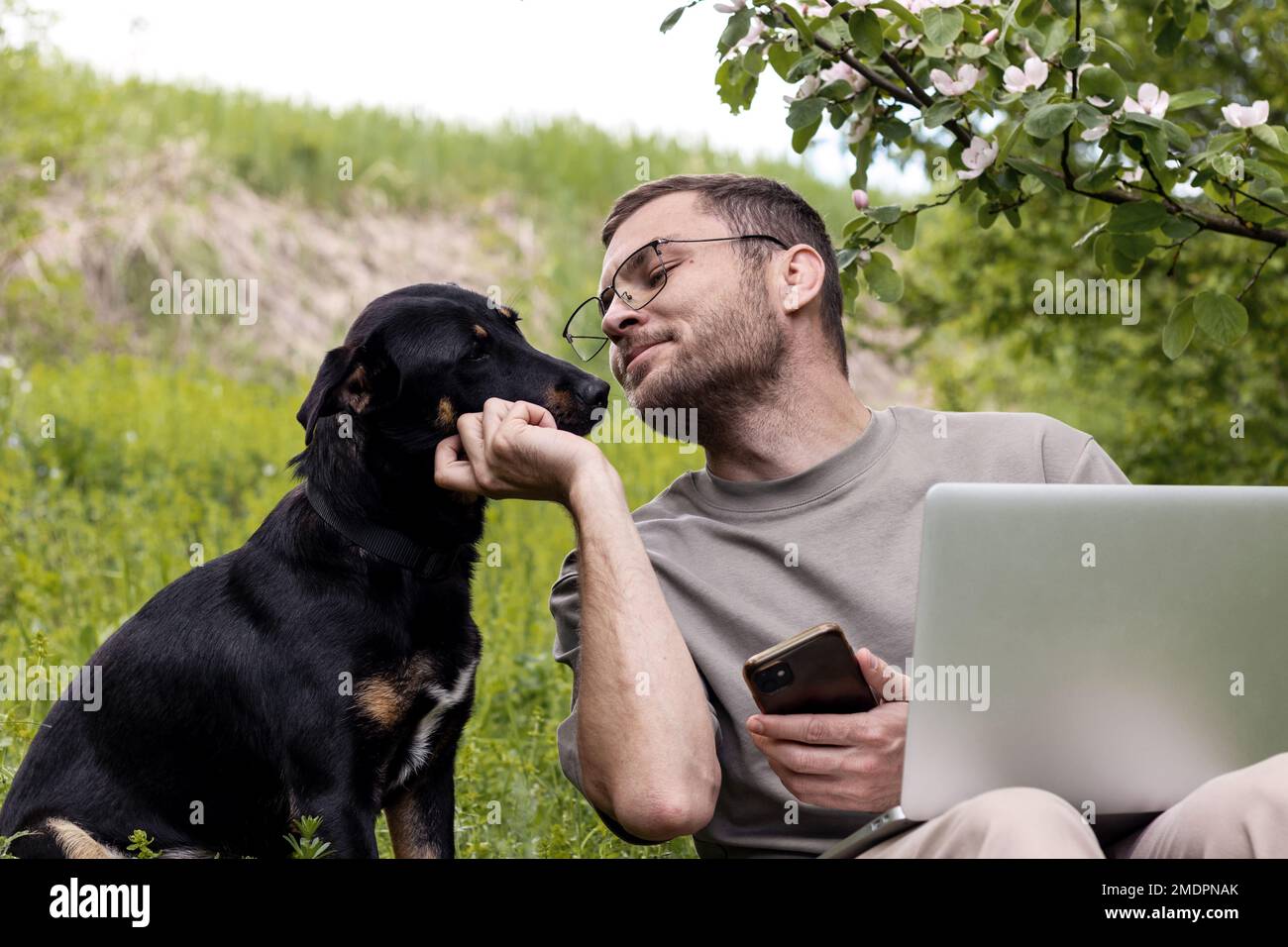 Uomo proprietario di un animale domestico che lavora su un computer portatile all'aperto e tocca delicatamente la faccia del suo cane. Amore e tenerezza per gli animali domestici. Foto Stock