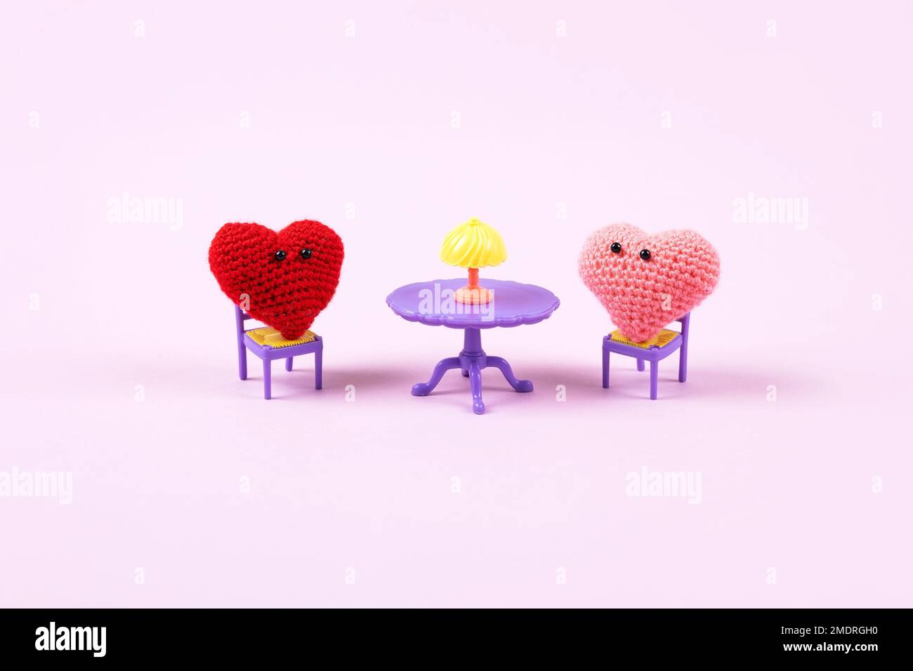 Concetto di San Valentino. Composizione di due cuori rossi e rosa lavorati a maglia seduti su sedie giocattolo su un tavolo giocattolo viola su sfondo lilla. Simbolo o Foto Stock